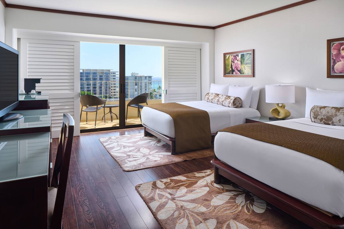 A hotel room with two beds, a TV, a desk, and a balcony with a view of the city.