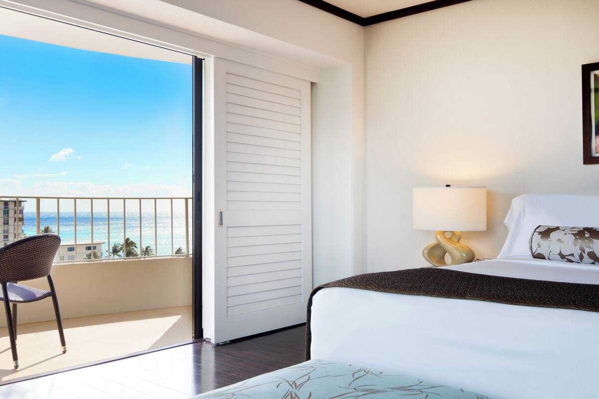 A hotel room with a bed, a chair, a lamp, and a view of the ocean.