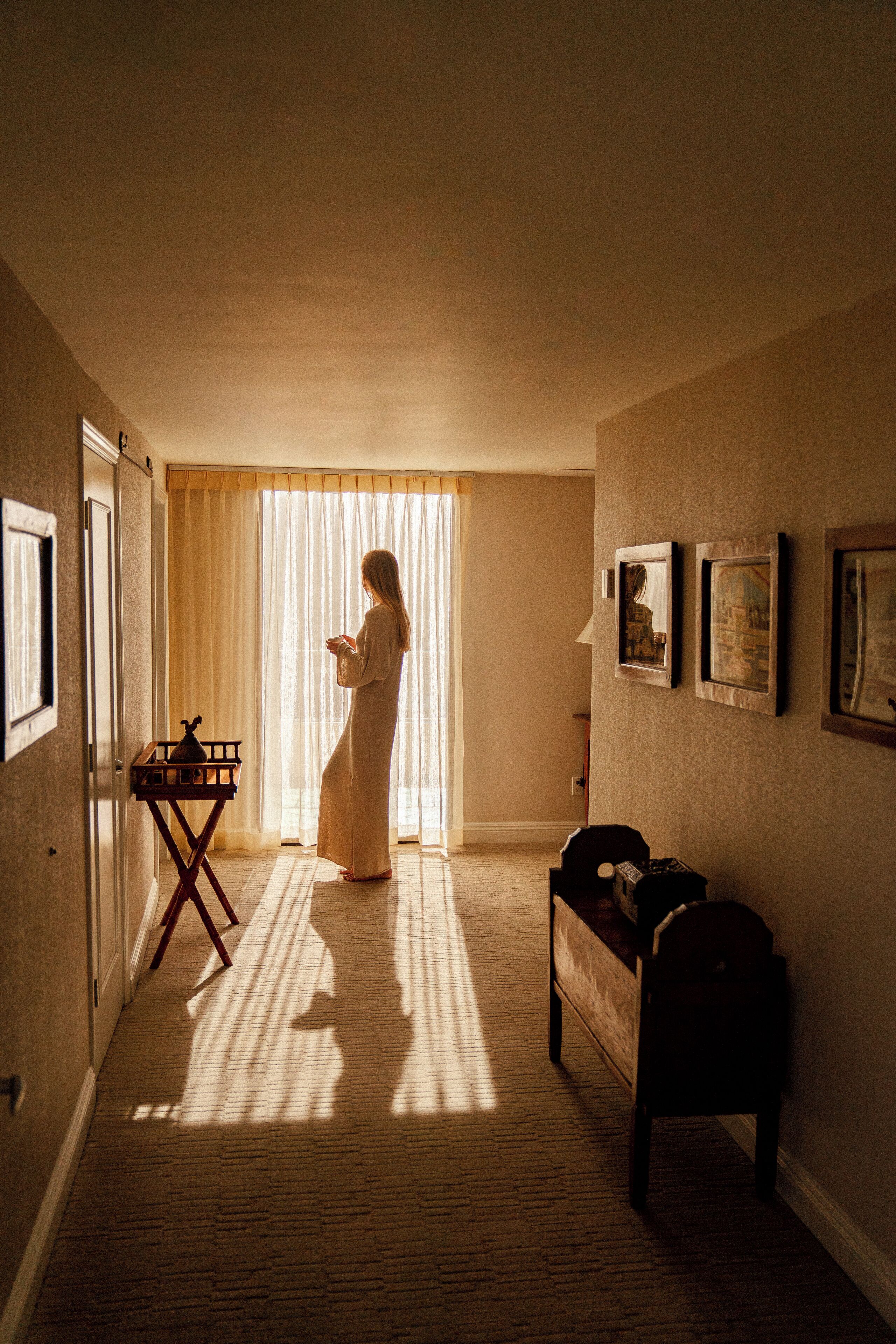 Een hotelgang met een vrouw die bij een raam staat, een klein tafeltje met een waterkoker, en ingelijste foto's aan de muur.