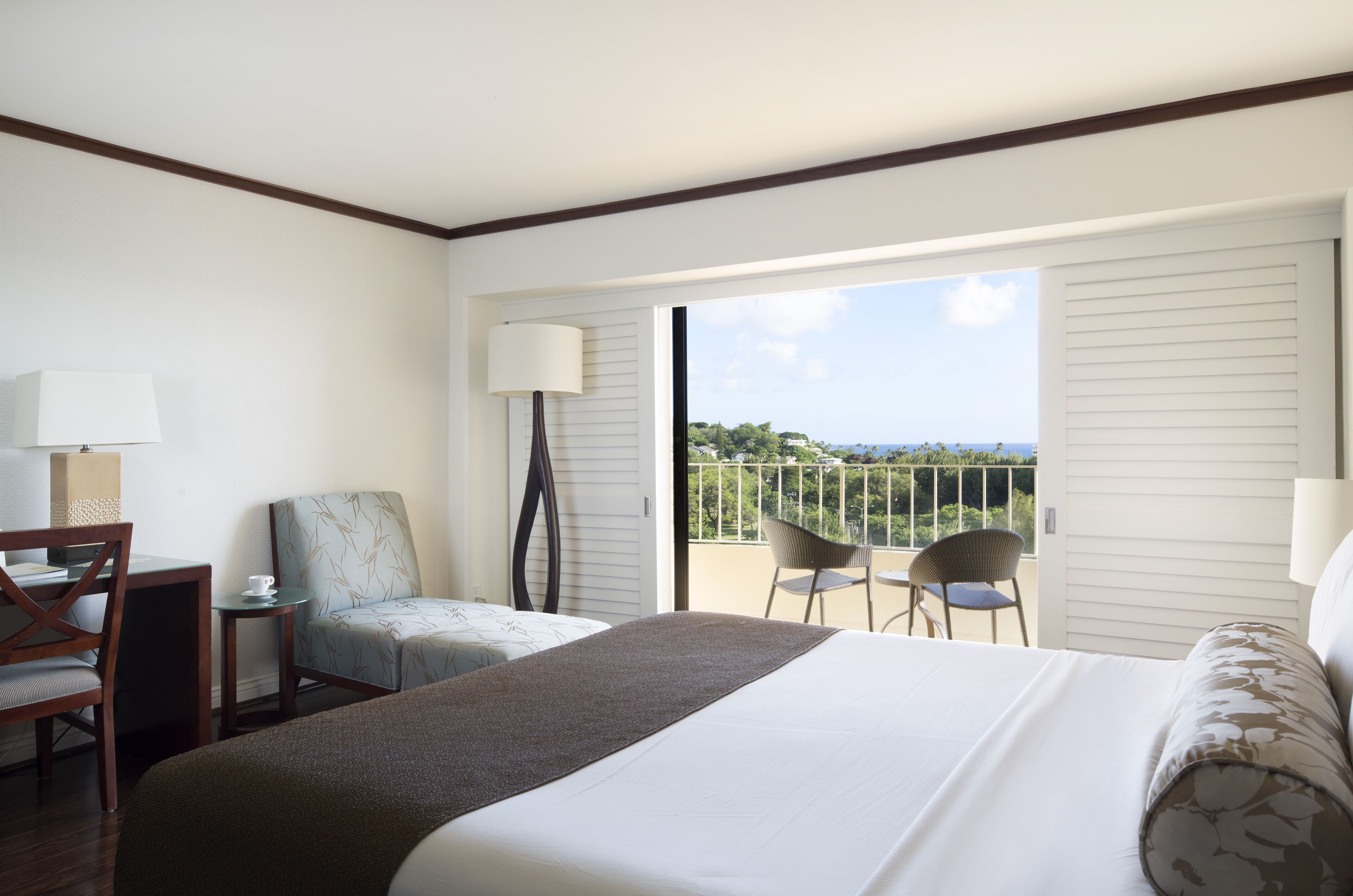 Une chambre d’hôtel avec un lit, un bureau avec une lampe, deux chaises et un balcon avec vue.