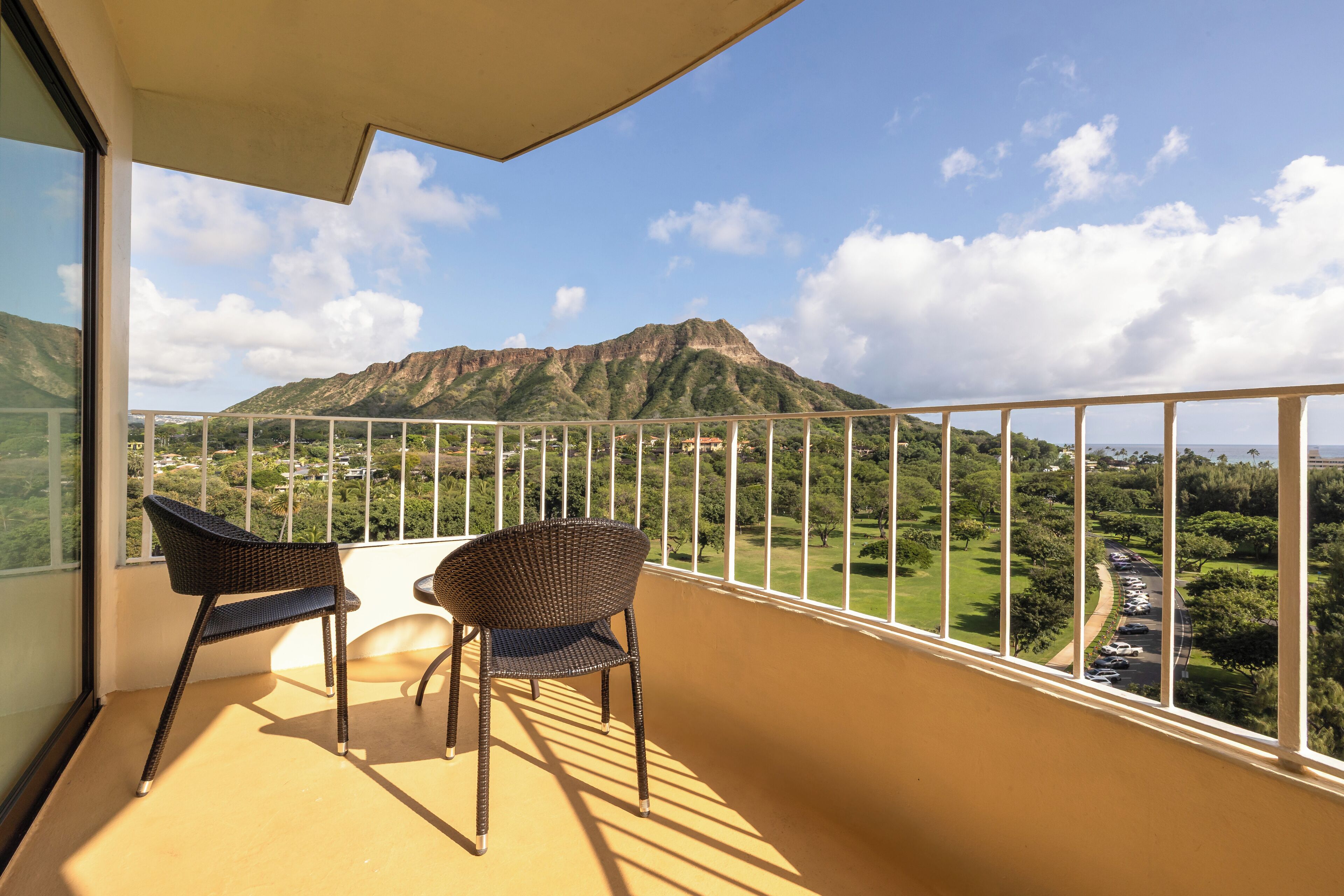 Un balcon avec deux fauteuils en osier, une vue sur une montagne et un espace de stationnement.
