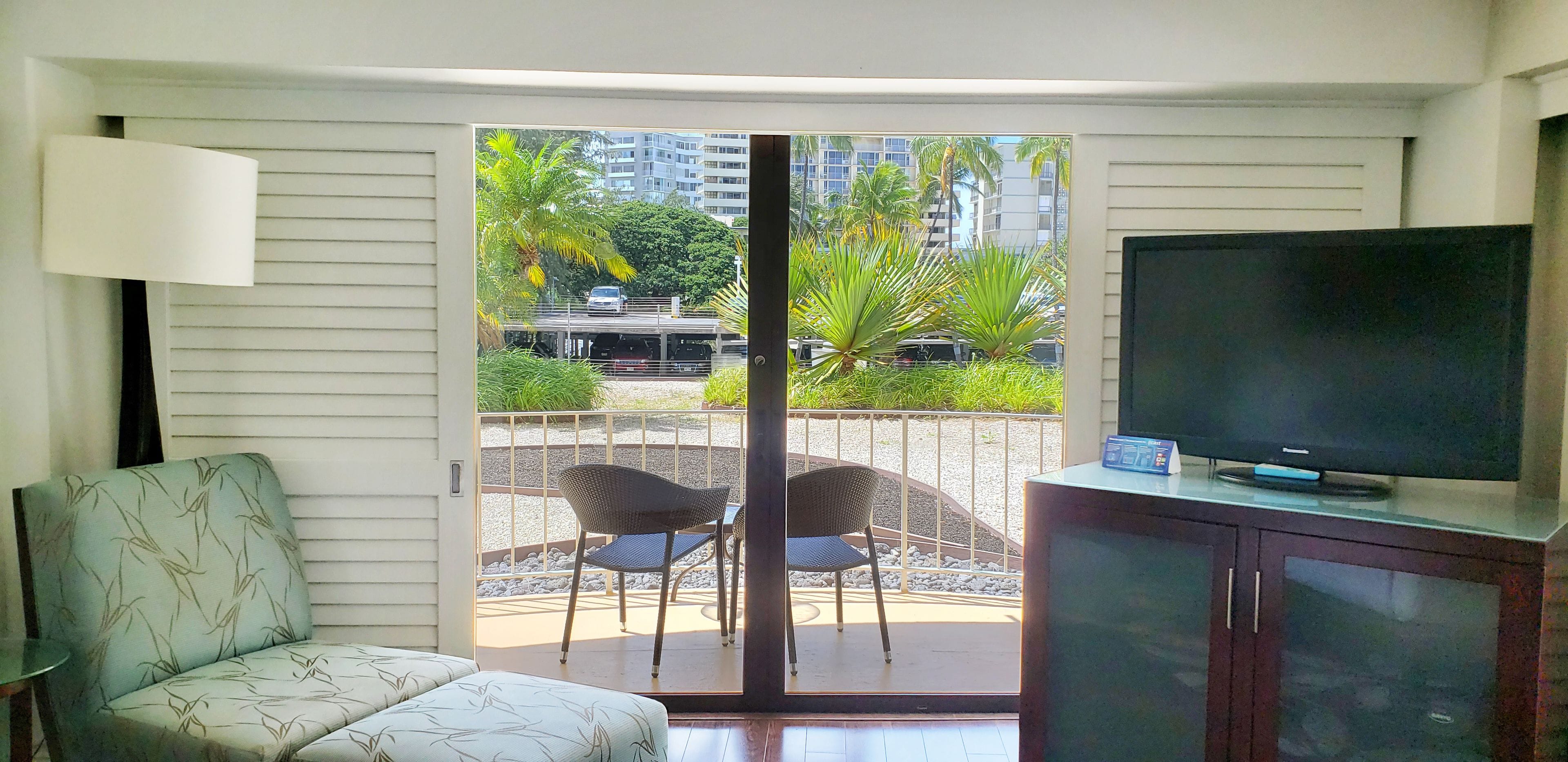 Una habitación con sofá, televisor sobre un mueble bajo y puerta corrediza de vidrio que da a un espacio exterior con sillas y vistas a edificios y palmeras.