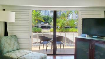 Una habitación con sofá, televisor sobre un mueble bajo y puerta corrediza de vidrio que da a un espacio exterior con sillas y vistas a edificios y palmeras.
