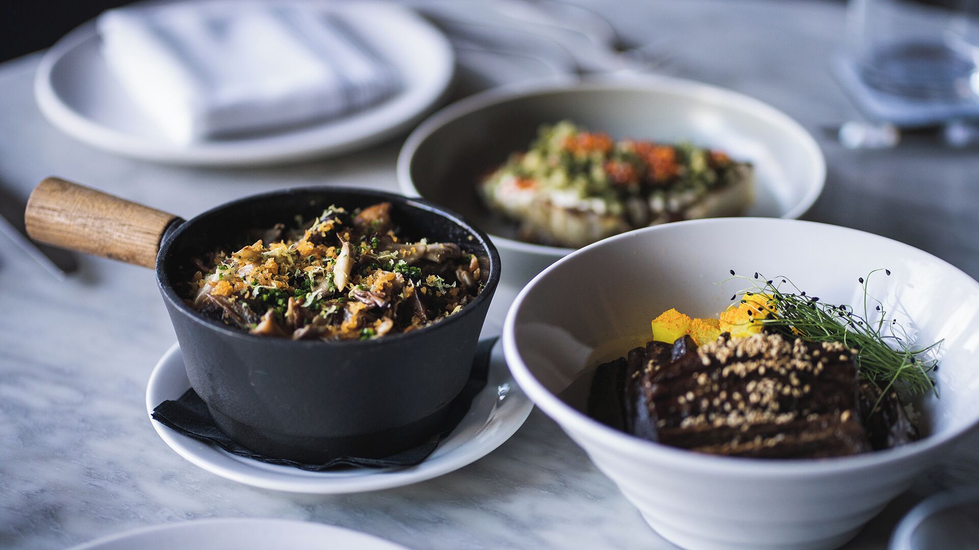 A table with three bowls of food, a wooden-handled pan, and a white plate with a napkin.