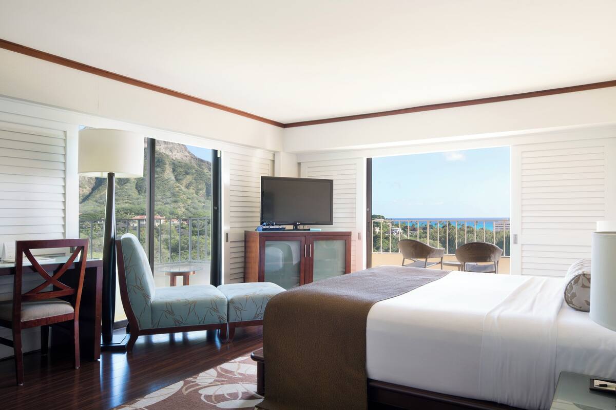 A hotel room with a bed, a TV, a desk, and a chair. There is a balcony with a view of the ocean.