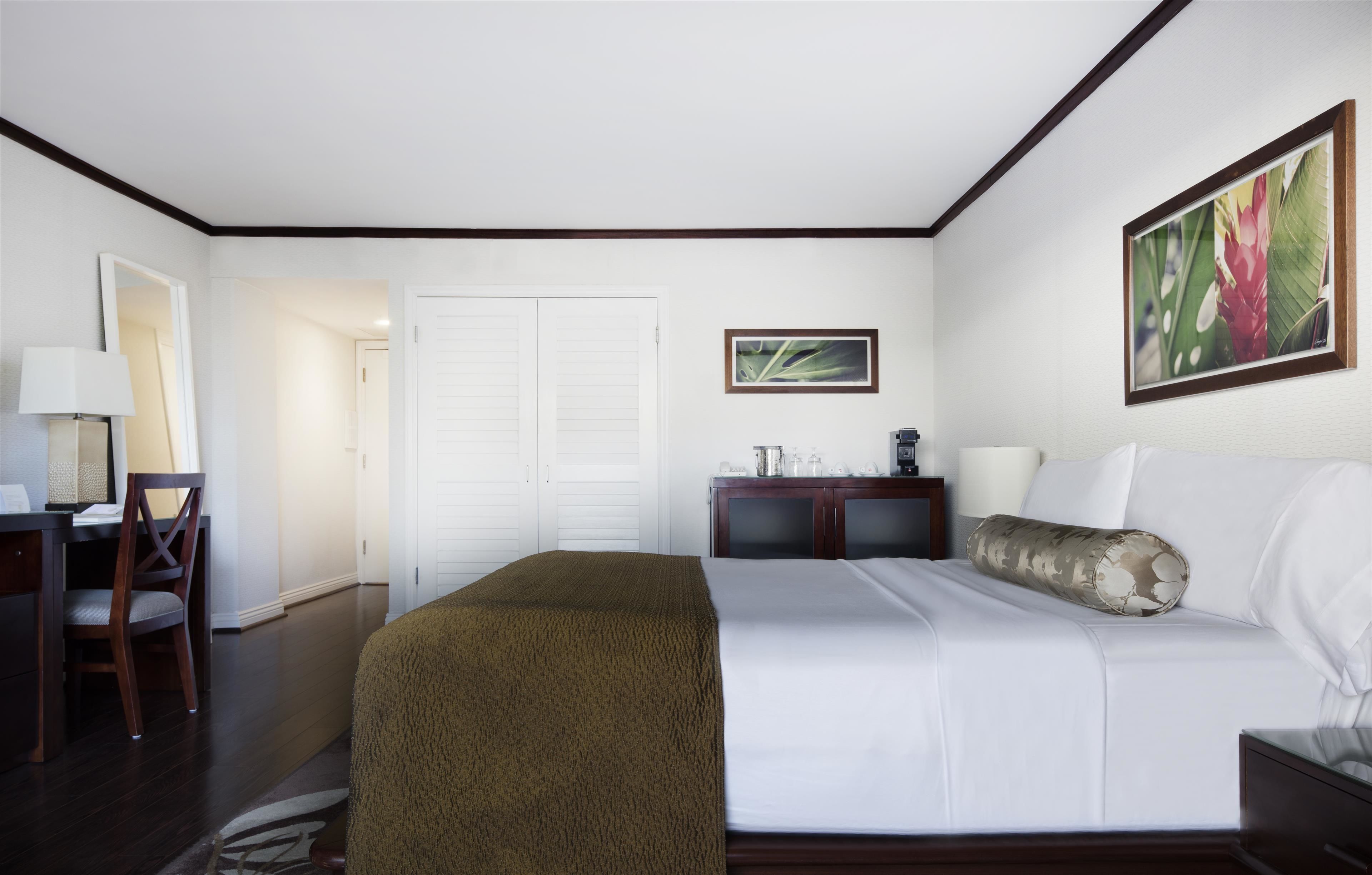 A hotel room with a bed, a desk with a chair, a wardrobe, and a painting on the wall.