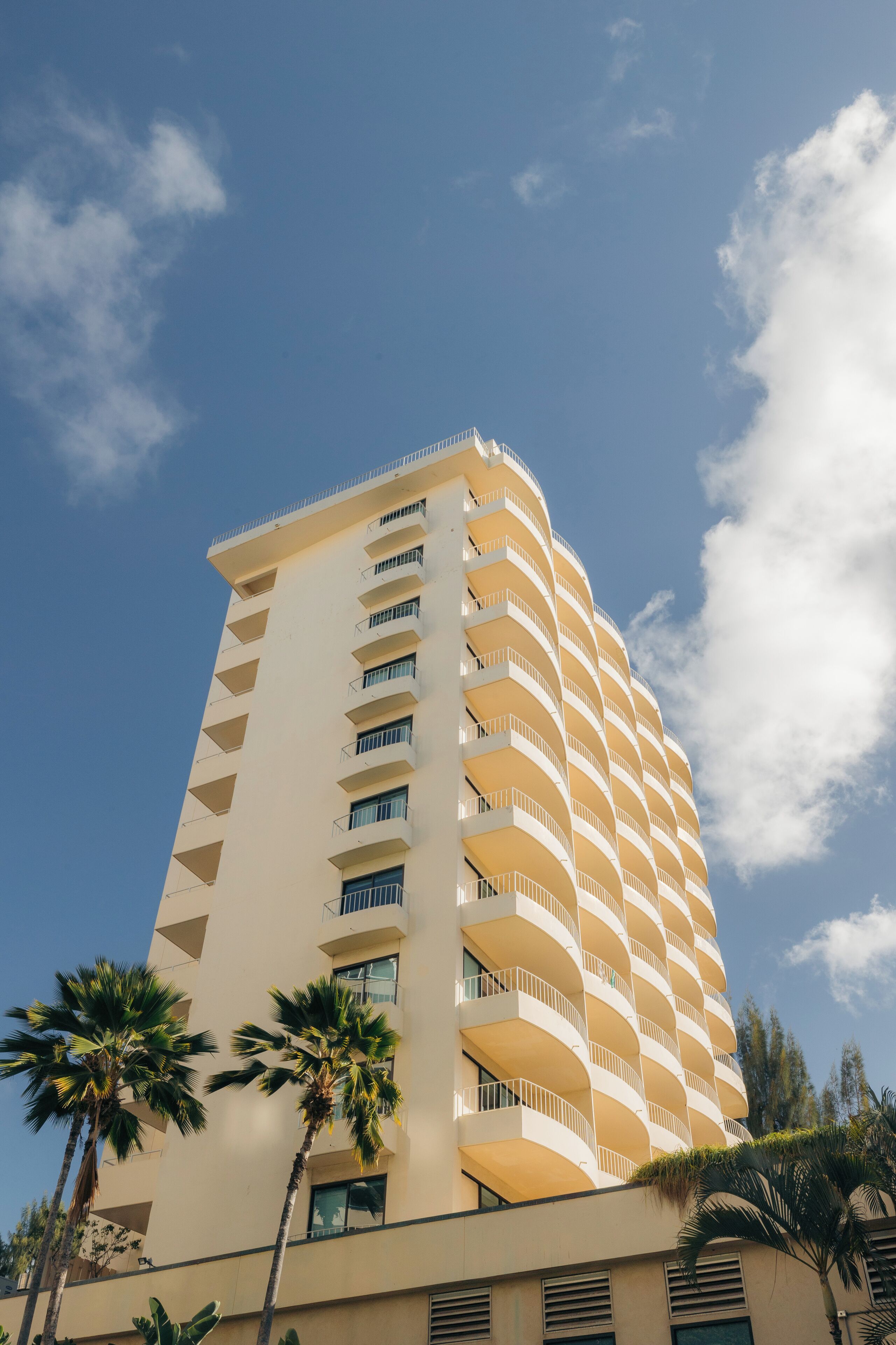Un edificio alto de varios pisos, de color beige y con balcones, palmeras en primer plano y un cielo azul despejado con nubes.
