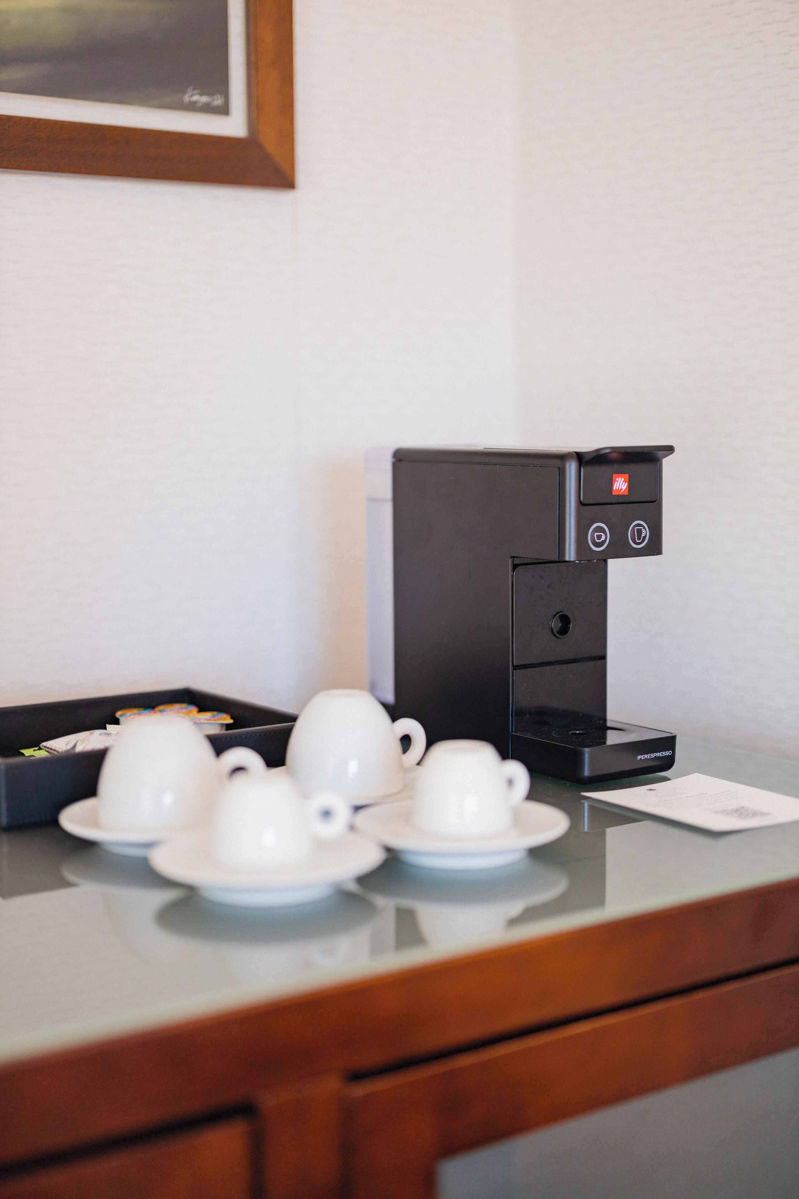 Eine Kaffeemaschine auf einem Tisch, dazu weiße Tassen und Untertassen, eine Kapselauflage und ein gerahmtes Bild an der Wand.