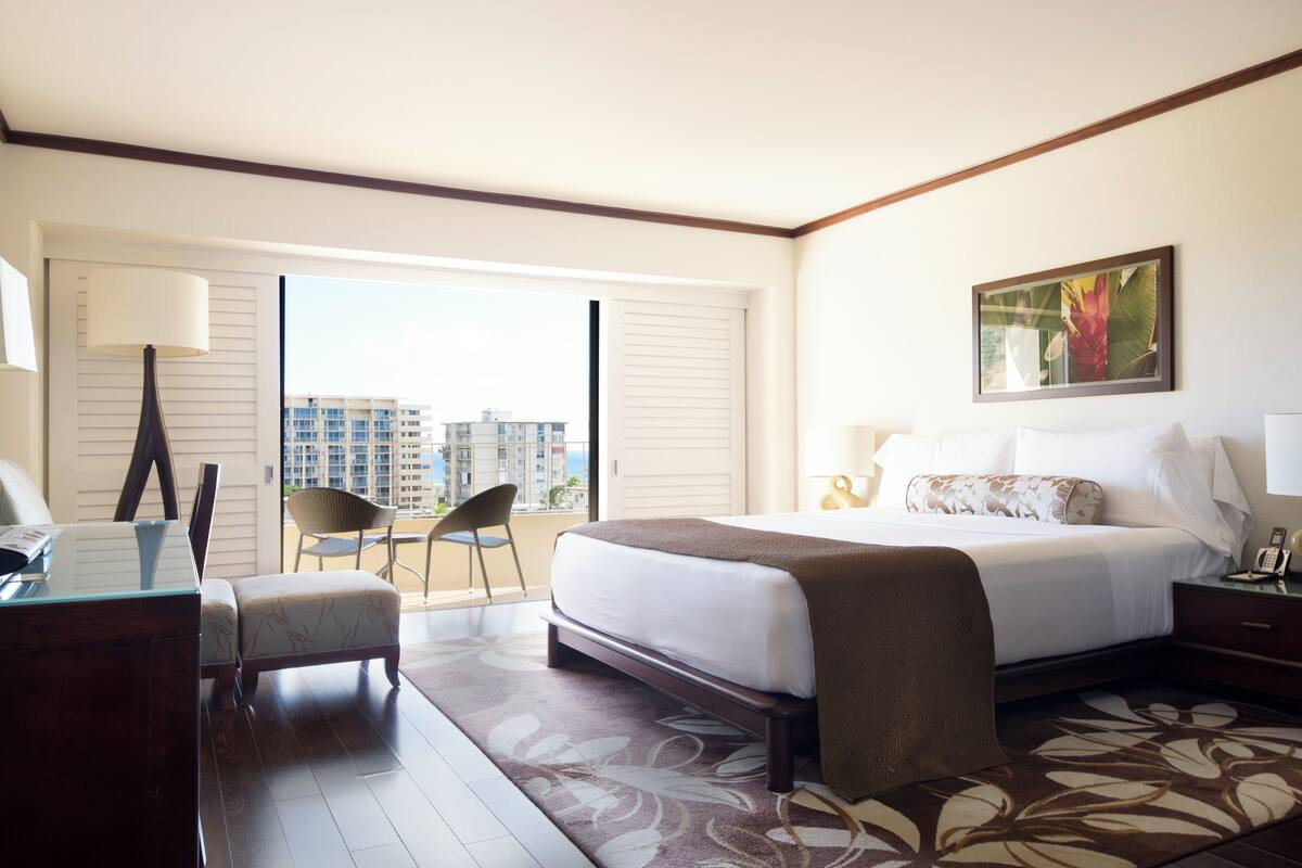 A hotel room with a large bed, a desk, a chair, and a view of buildings through the window.