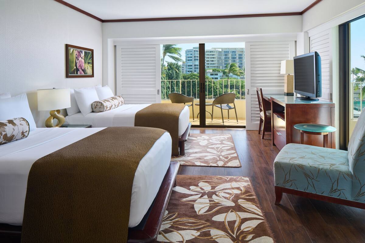 A hotel room with two beds, a desk with a TV, and a balcony with chairs.