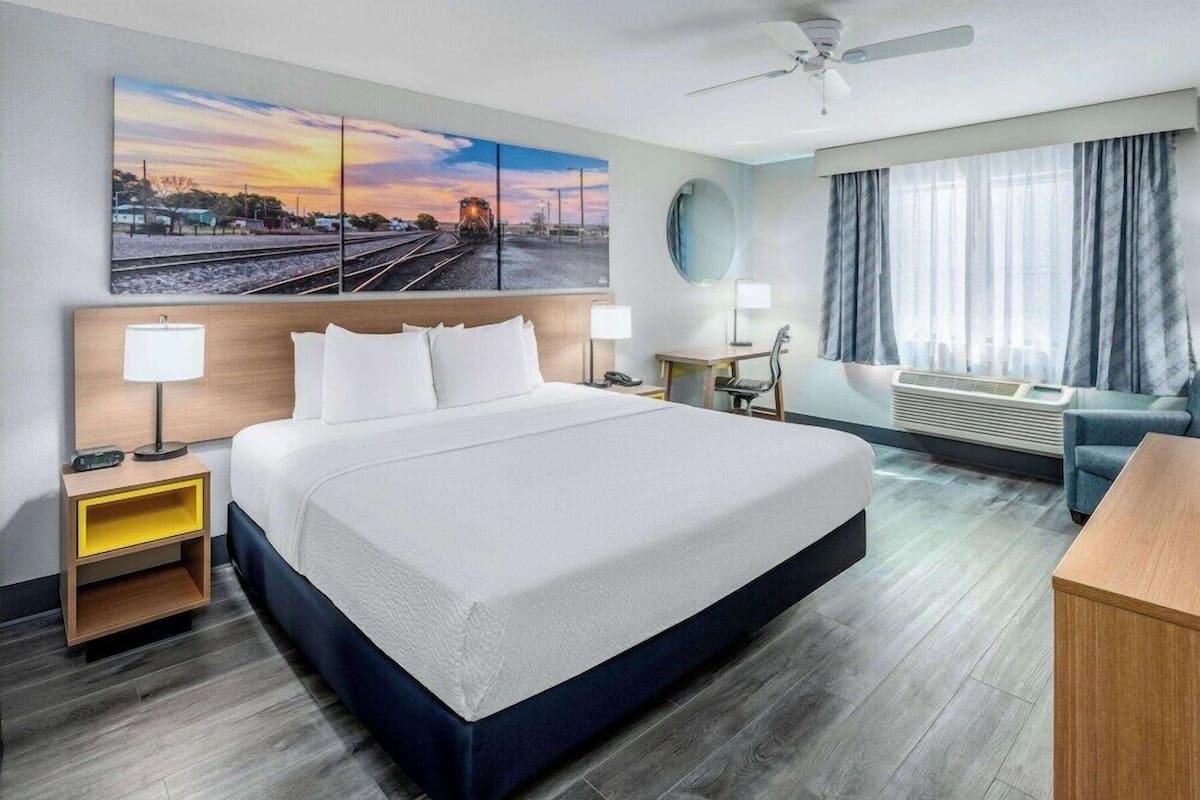 A hotel room with a large bed, bedside tables, a desk, a chair, a ceiling fan, a large window with curtains, and a wall-mounted TV.