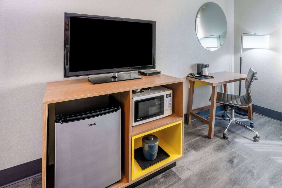 A compact hotel room with a flat-screen TV, a microwave, a mini-fridge, a desk with a chair, and a mirror.