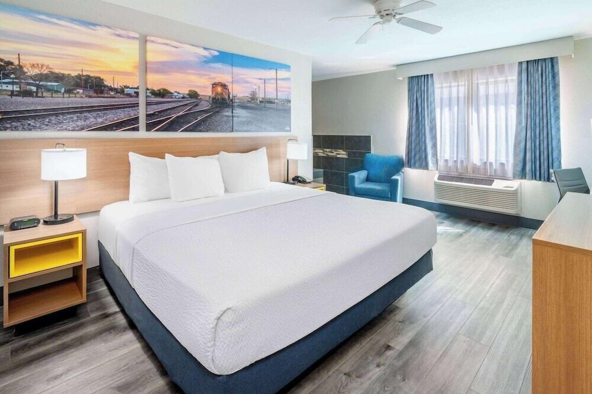 A hotel room with a large bed, a desk, a chair, a window with curtains, a ceiling fan, and a painting of a train on the wall.