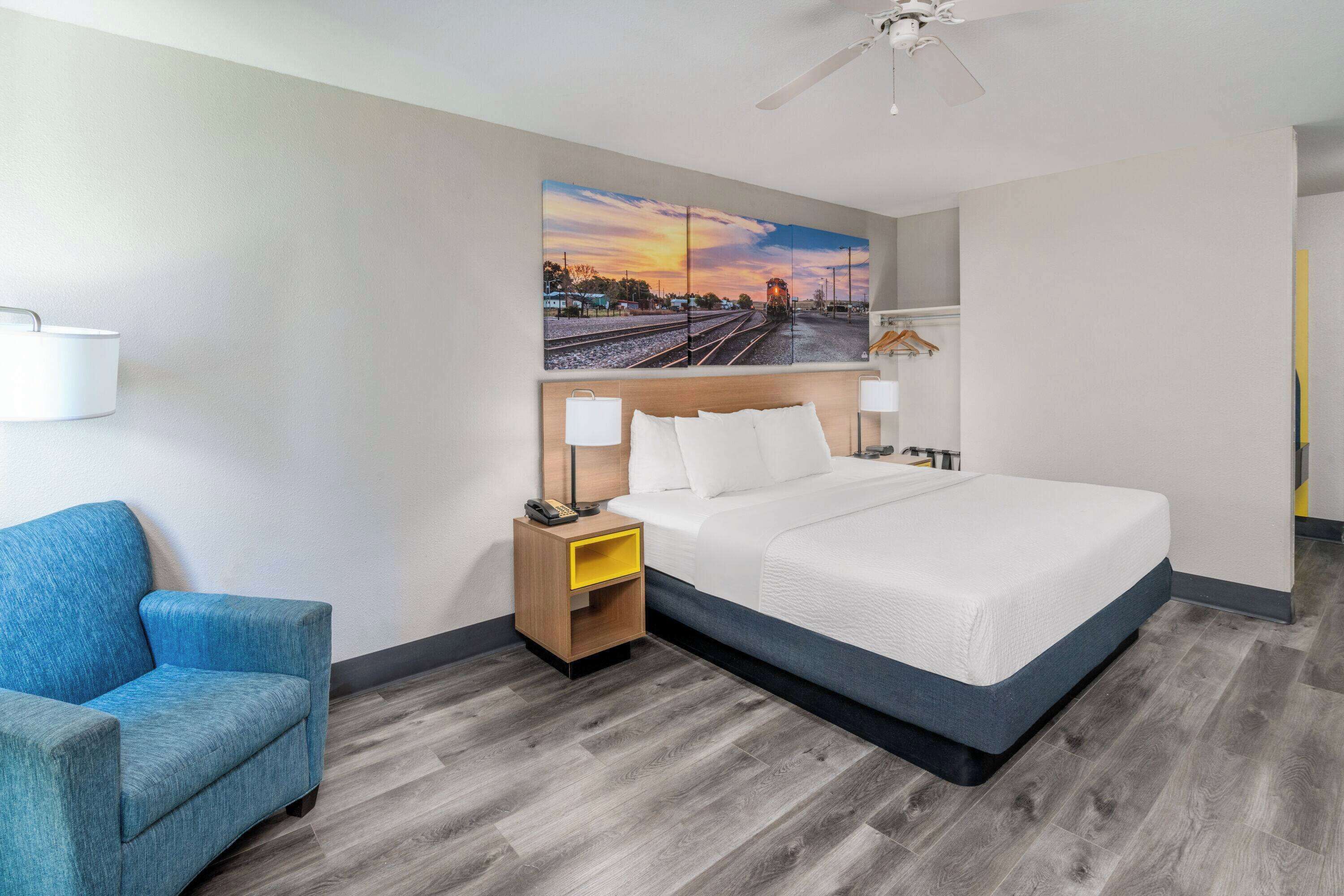 A hotel room with a large bed, a blue armchair, a nightstand with a lamp, and a painting of a sunset over a railway track.