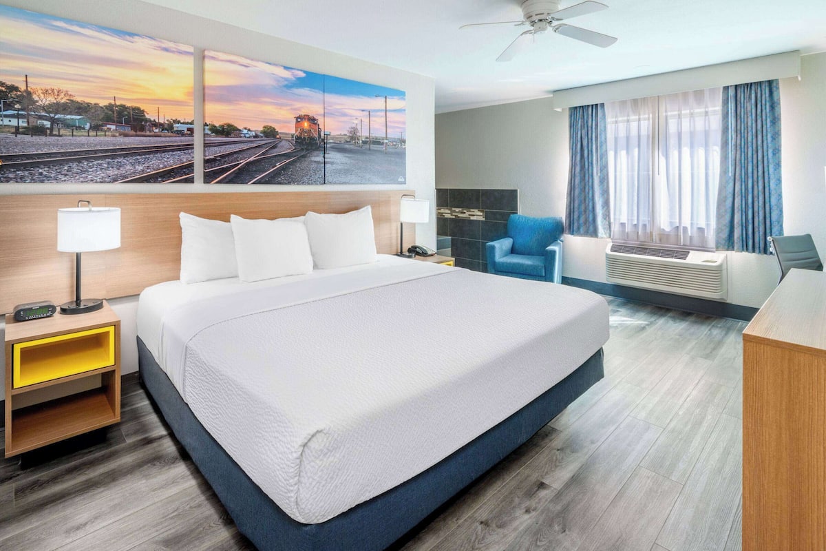 A hotel room with a large bed, a desk, a chair, a nightstand, a lamp, a painting of a train on the wall, and a ceiling fan.