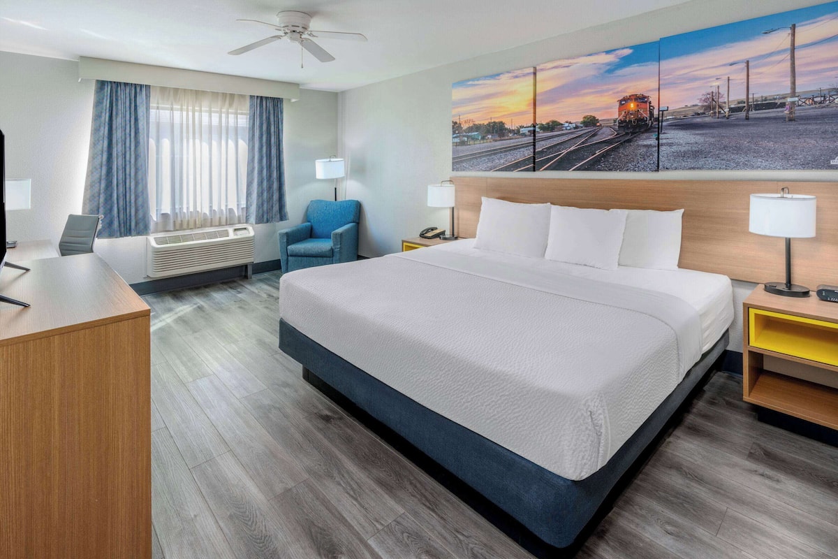 A hotel room with a large bed, a desk, a chair, a desk lamp, a nightstand, and a painting of a train on the wall.