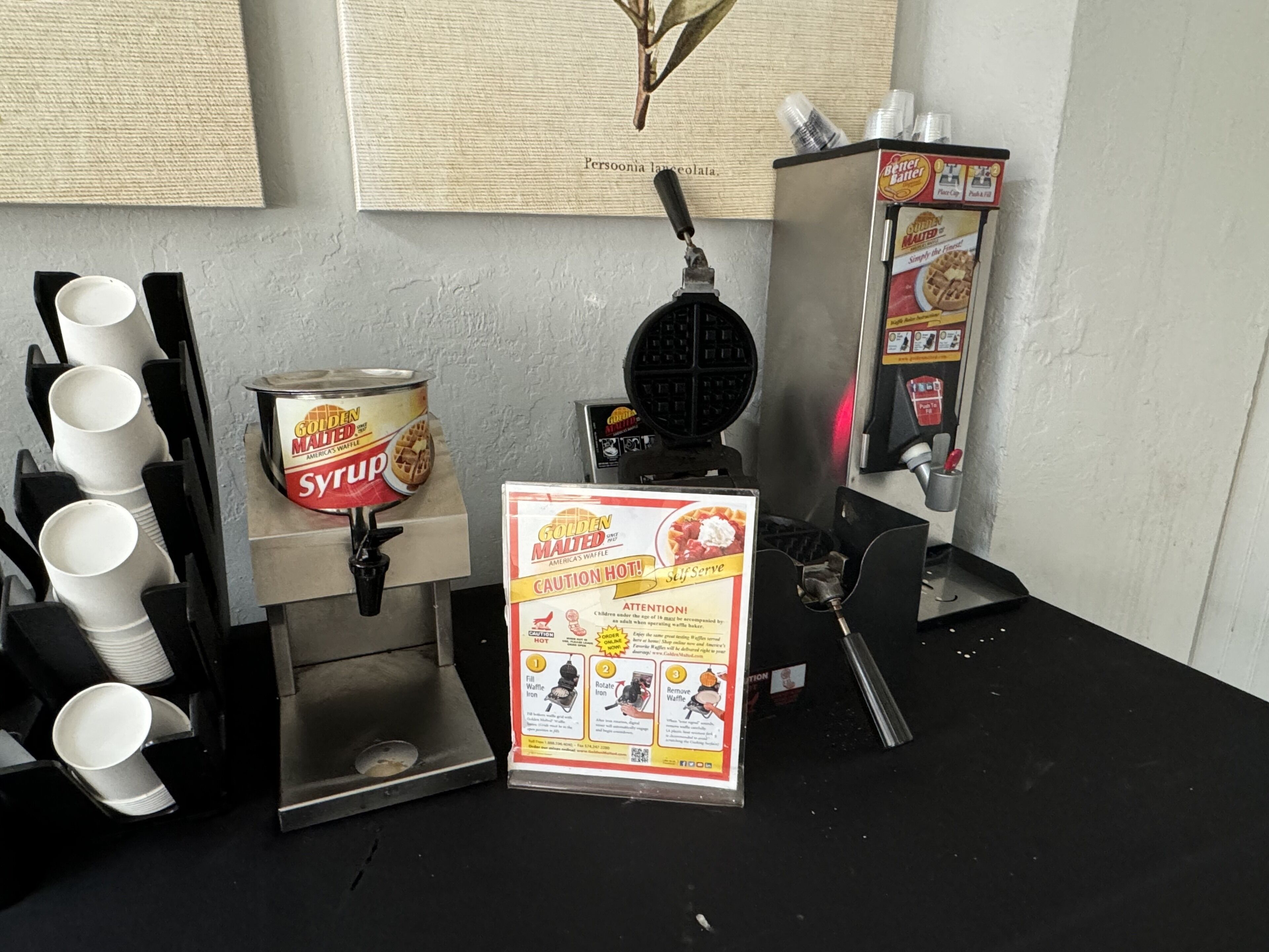 A self-serve waffle station with syrup and waffle iron dispensers, a caution sign, and a stack of white cups.