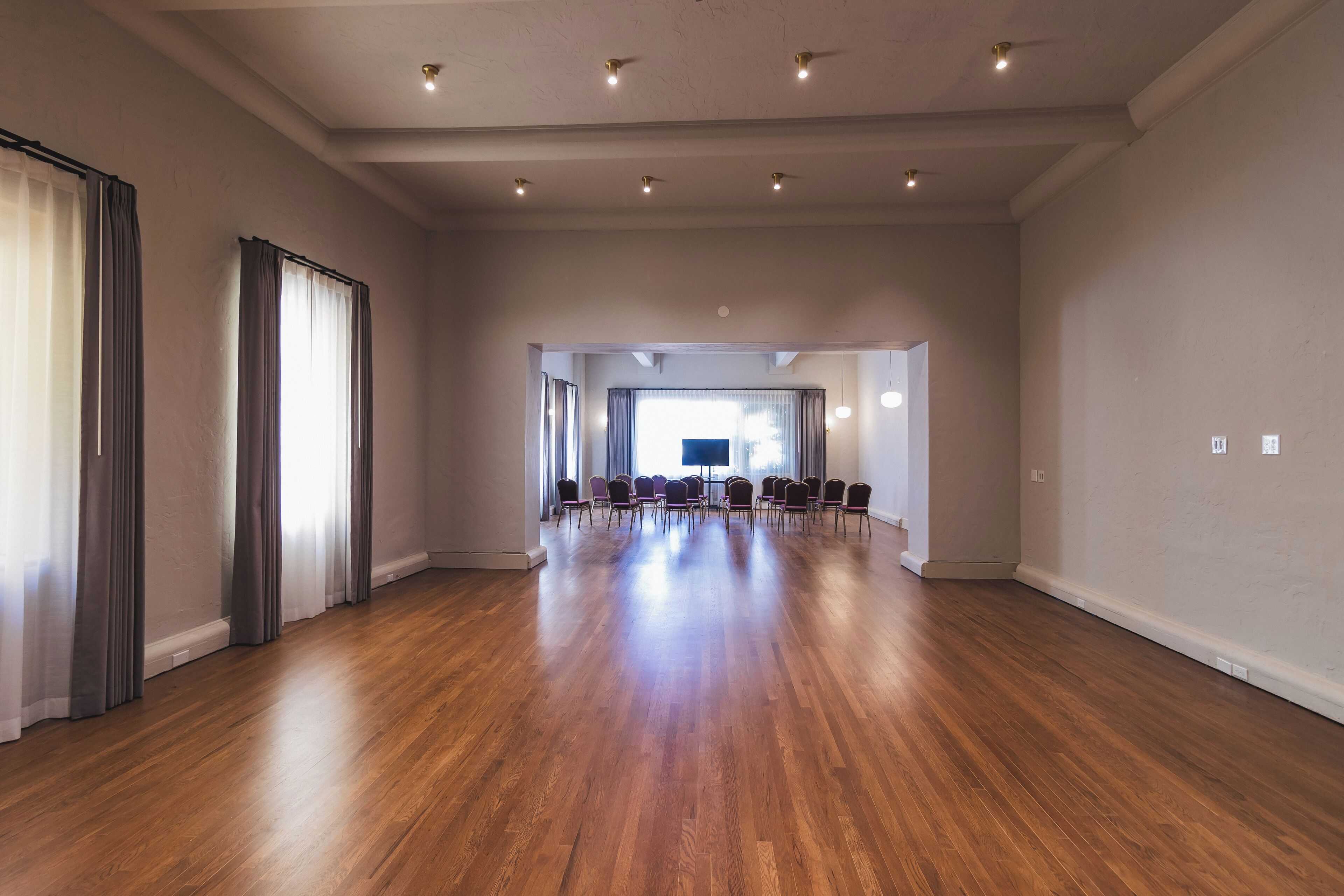 Una habitación amplia con suelo de madera, ventanas grandes con cortinas y una mesa de conferencias con sillas.