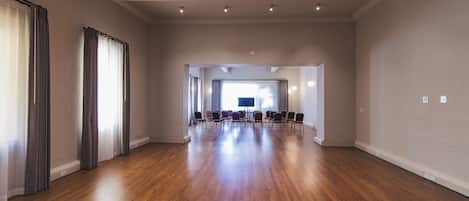 A spacious room with a wooden floor, large windows with curtains, and a conference table with chairs.