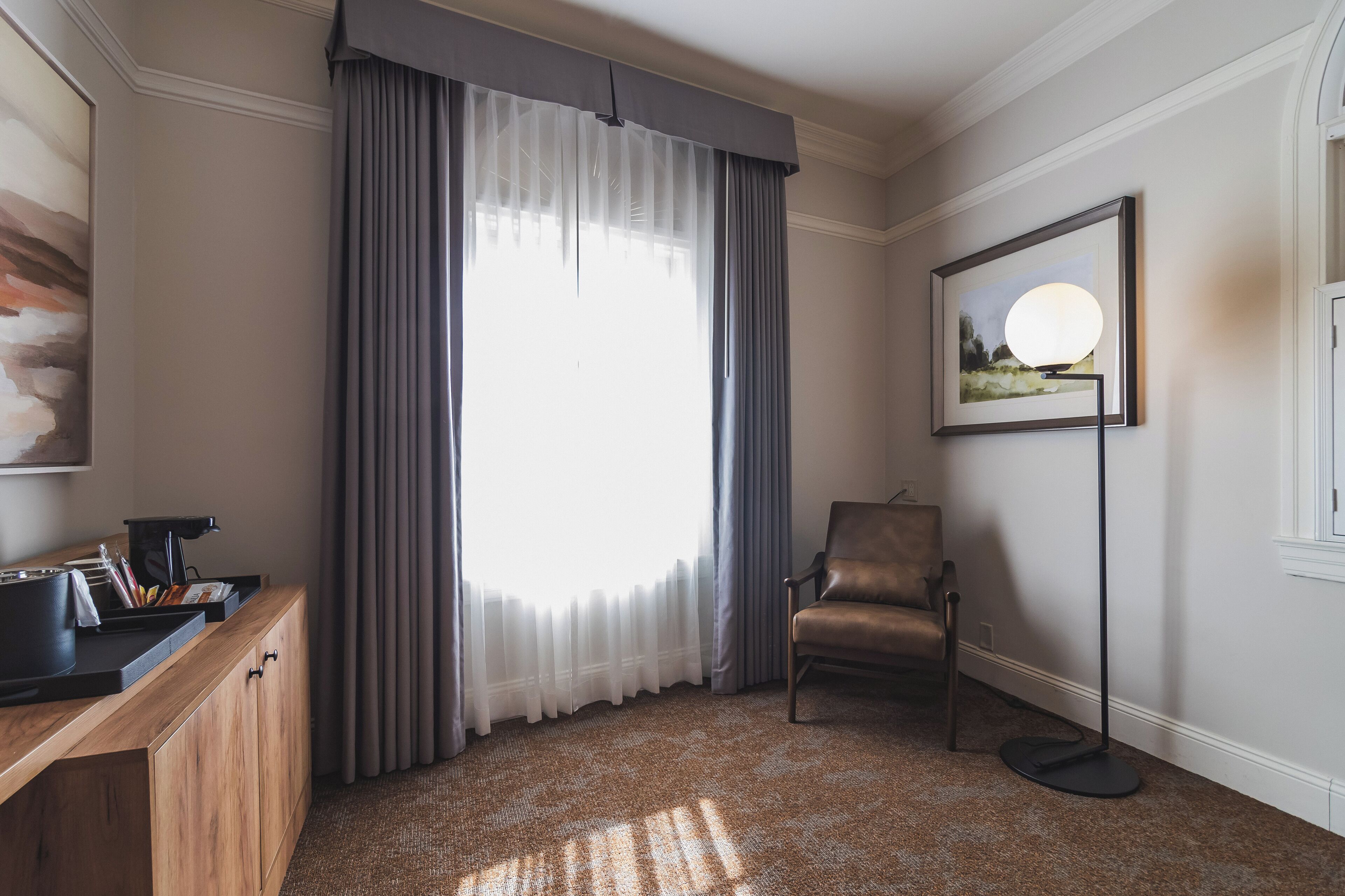 A hotel room with a wooden desk, a chair, a floor lamp, a large window with sheer curtains, and a framed picture on the wall.