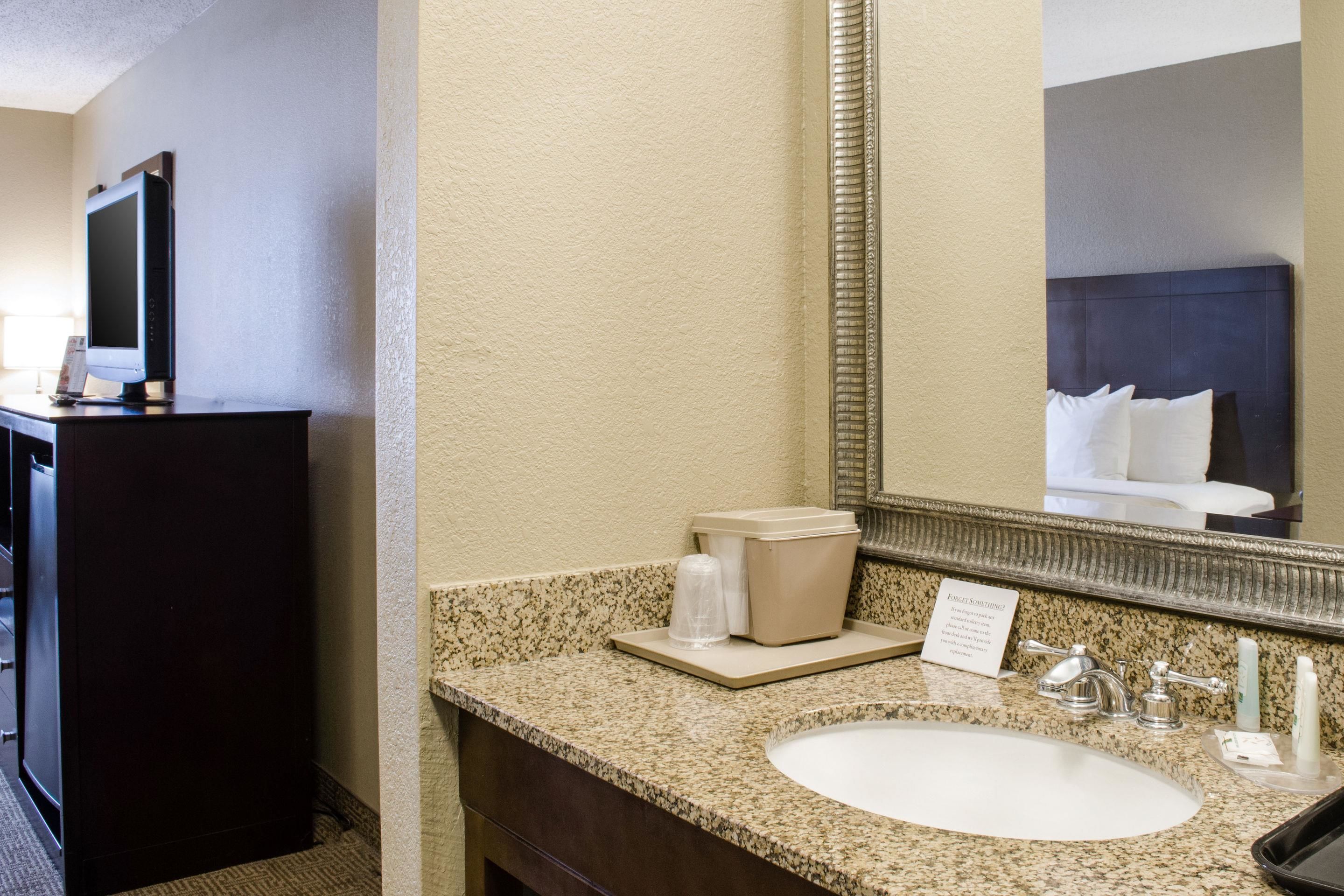 A hotel bathroom with a granite countertop, a sink, a mirror, a toilet, and a television on a cabinet.