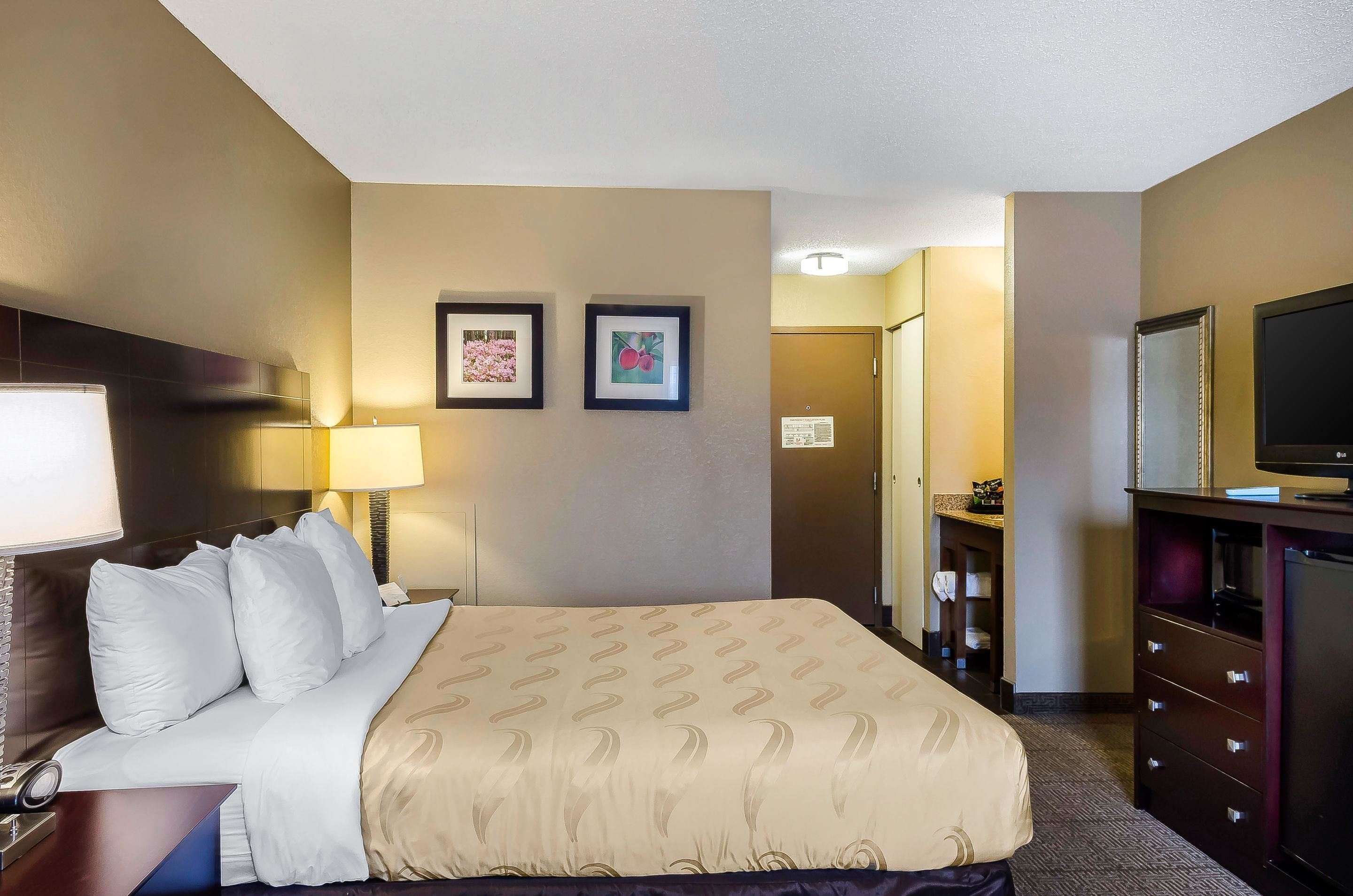 A hotel room with a bed, a desk, a lamp, a TV, and two framed pictures on the wall.