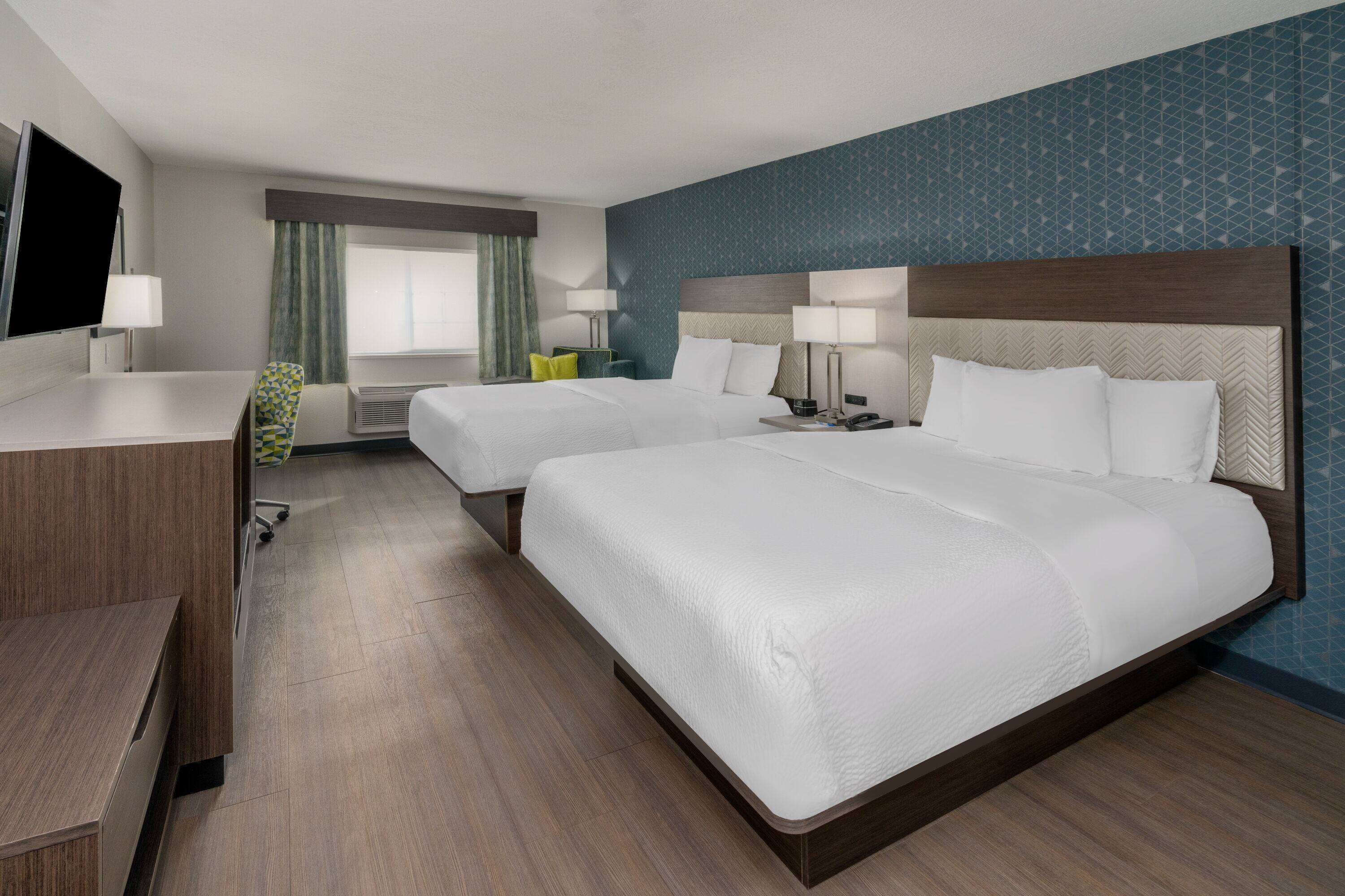 A hotel room with two beds, a desk, a TV, and a window with curtains.