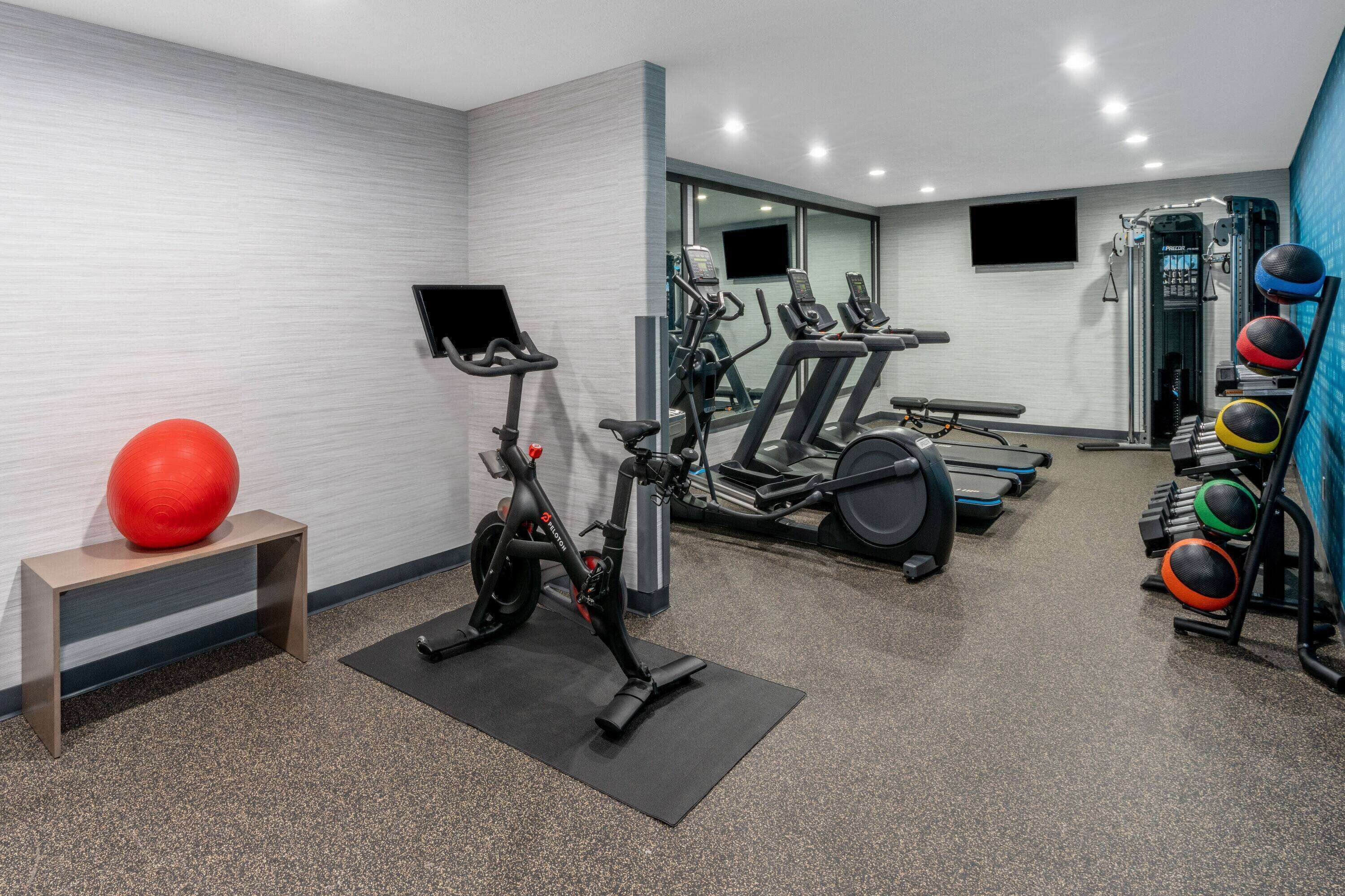 Un gimnasio con equipo de ejercicio, una pelota roja para hacer ejercicio y una televisión montada en la pared.