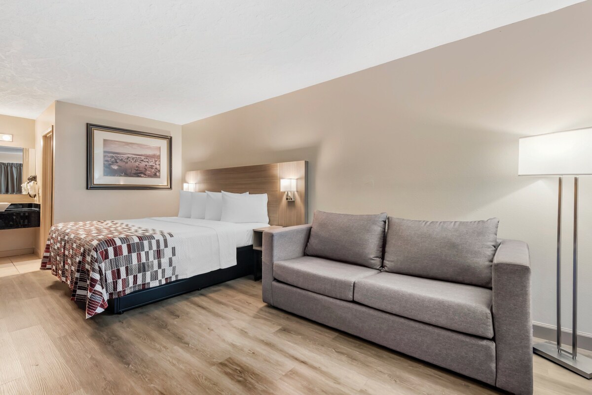 A hotel room with a bed, a grey sofa, a wooden floor, a framed picture on the wall, and a nightstand with a lamp.