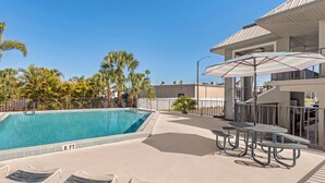 Ein Pool mit Liegestühlen und Sonnenschirmen, umgeben von Palmen und einem Gebäude mit einer überdachten Terrasse.