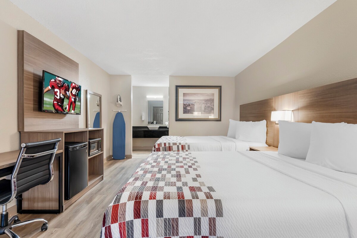 A hotel room with two beds, a desk with a chair, a television, a desk lamp, and a framed picture on the wall.