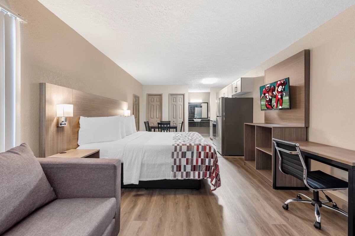 A hotel room with a bed, a desk, a chair, a TV, and a kitchenette.