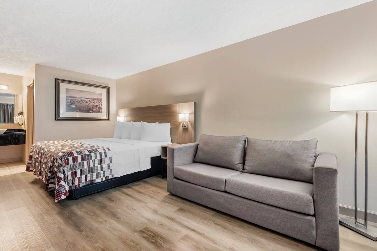 A hotel room with a bed, a grey sofa, a wooden floor, a framed picture on the wall, and a nightstand with a lamp.
