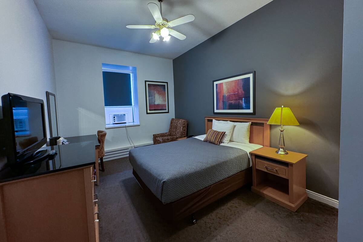 A hotel room with a bed, a desk with a television, a chair, a bedside table with a lamp, and artwork on the walls.