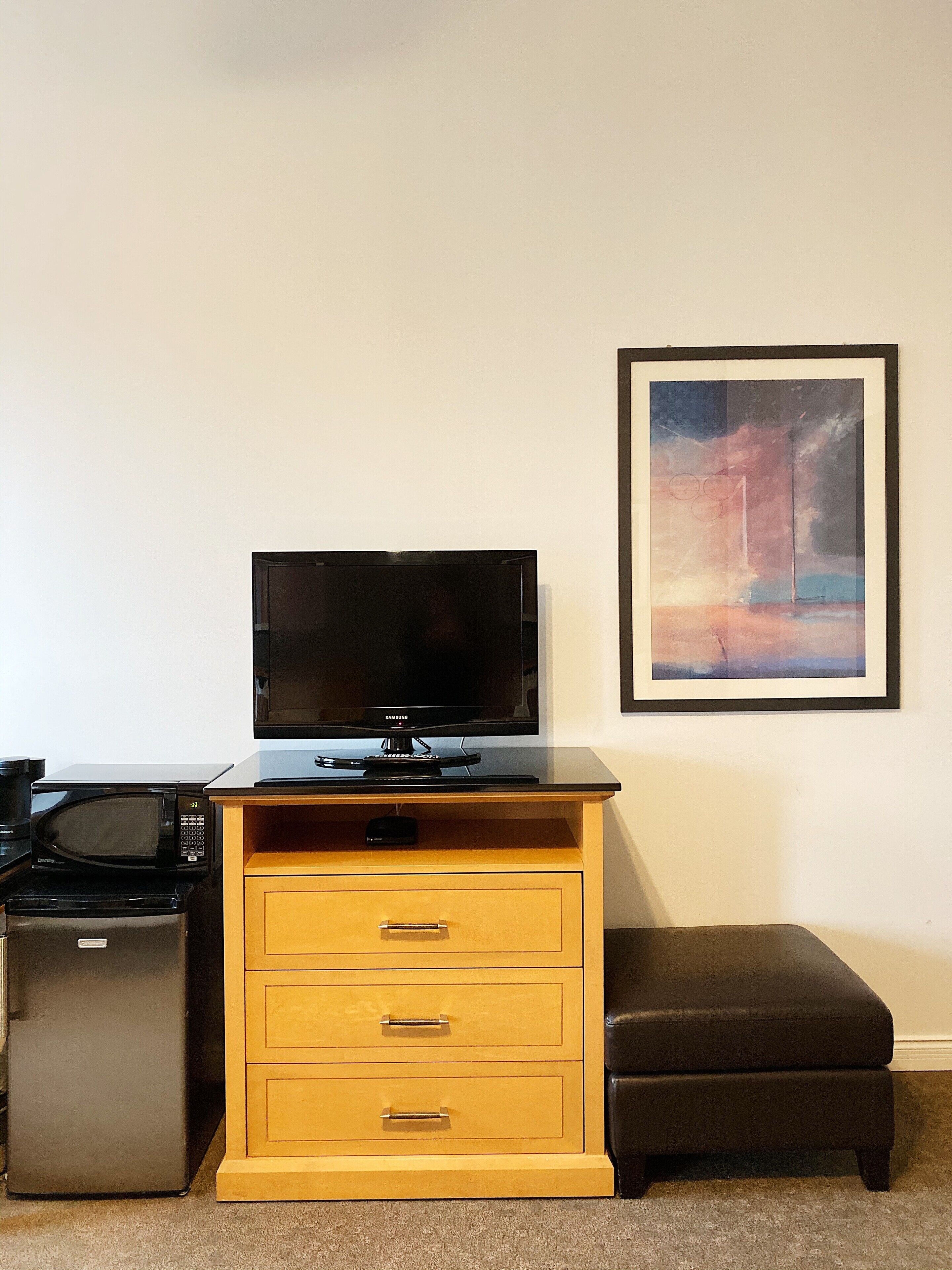 Una kitchenette con microondas, refrigerador y un televisor Samsung sobre un mueble de madera.