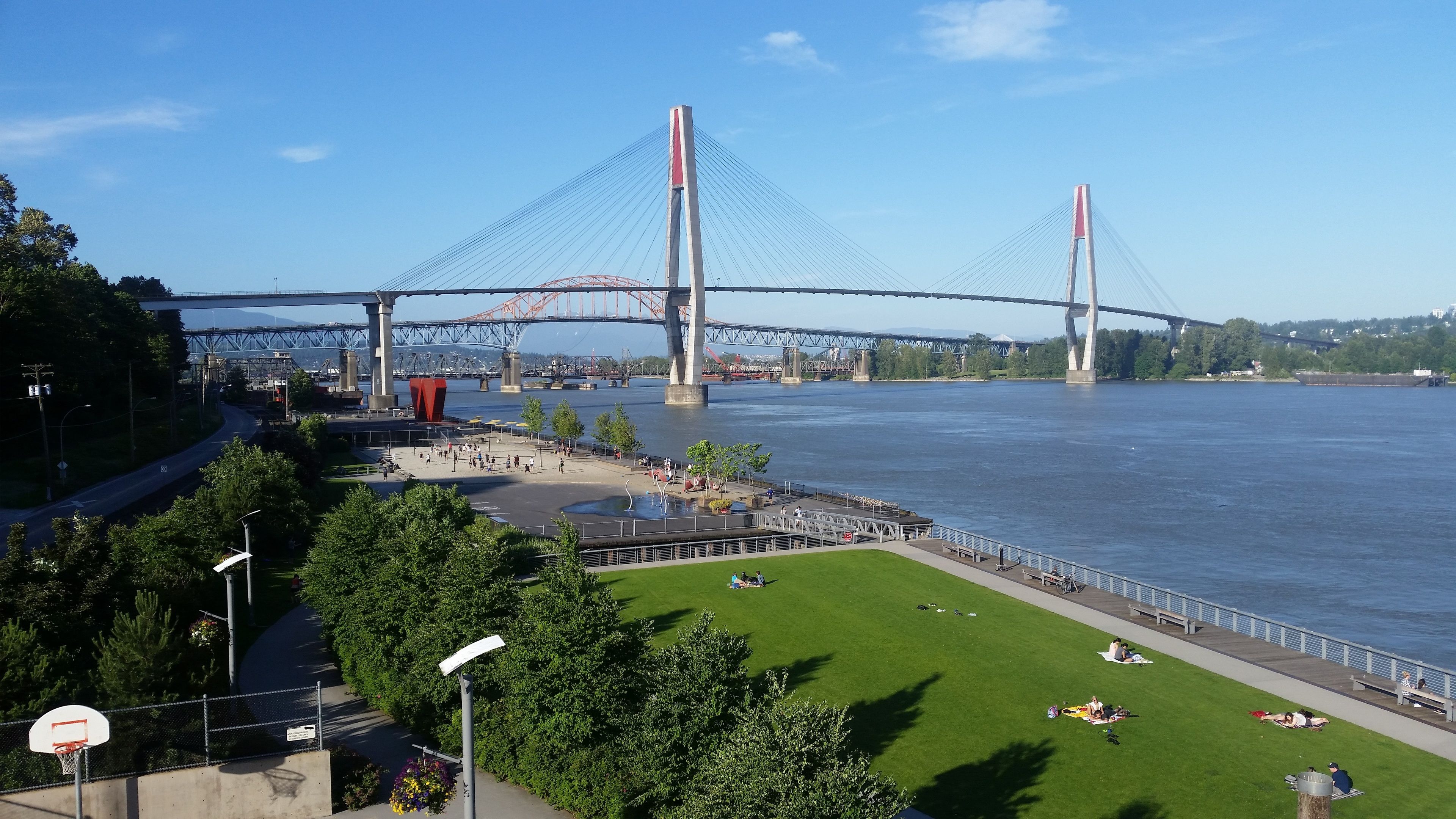 A riverside park with a basketball court, picnic areas, and a view of a large bridge.