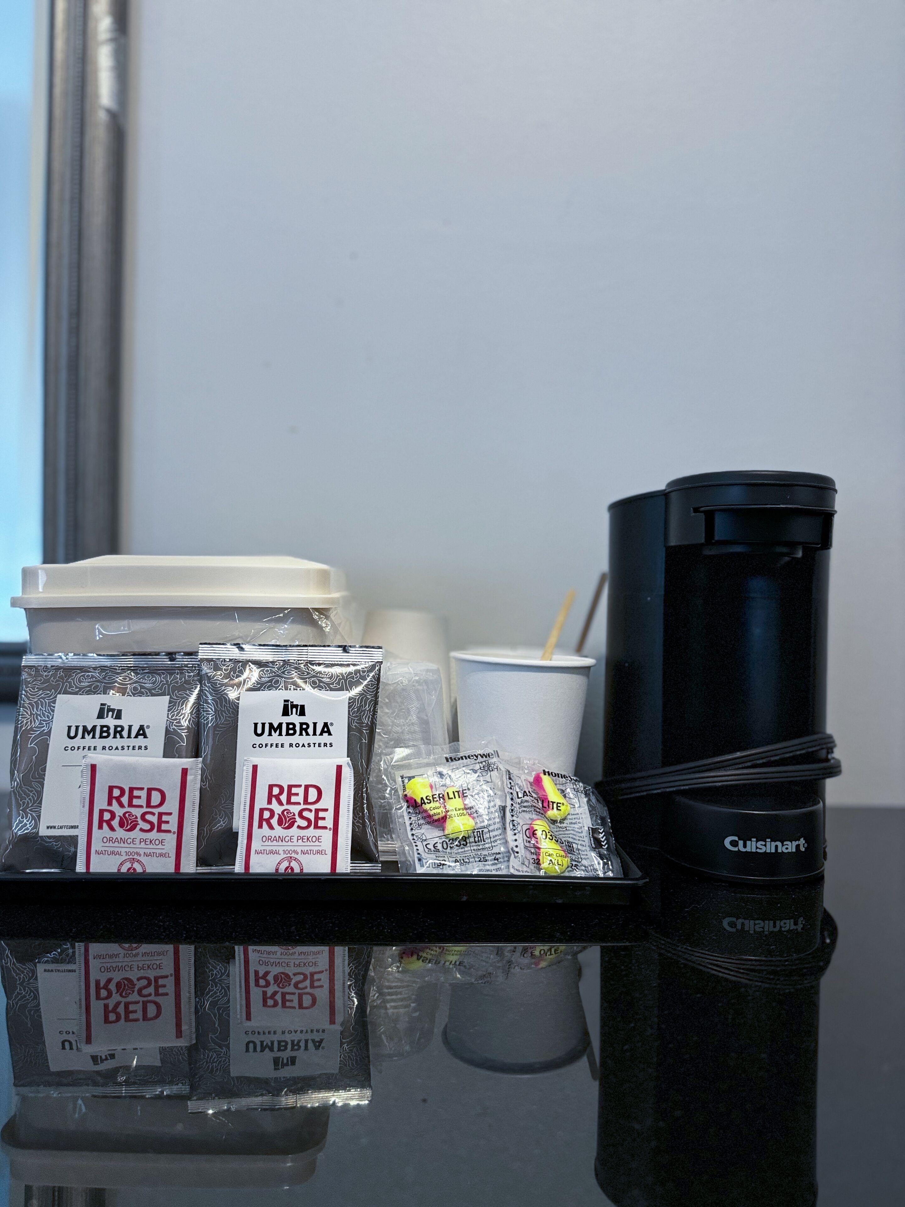 A coffee station with various coffee pods, a disposable cup, and a black coffee maker on a reflective surface.