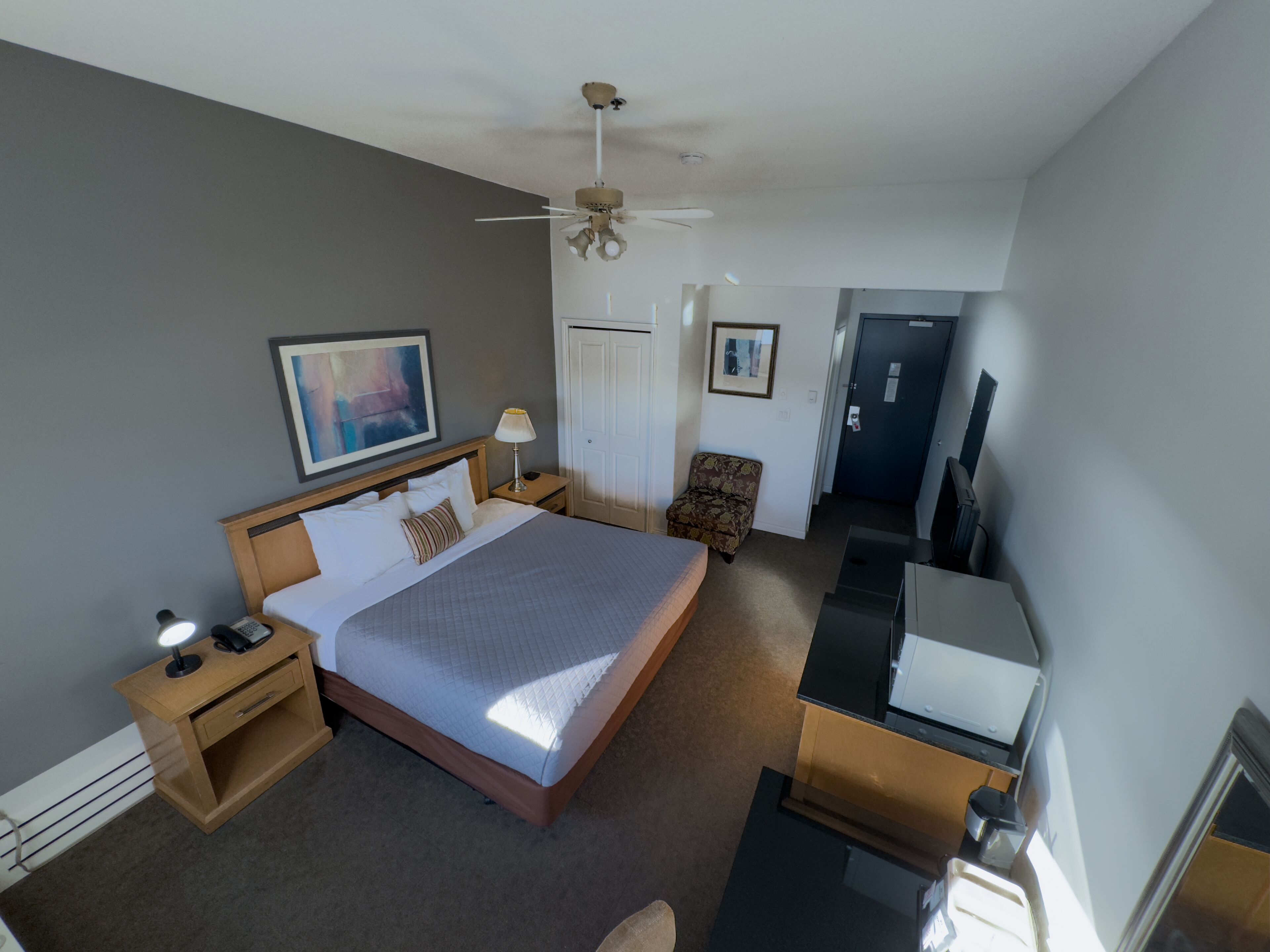 Une chambre d’hôtel avec un grand lit, un bureau avec un ordinateur, une chaise, un ventilateur de plafond et une porte.