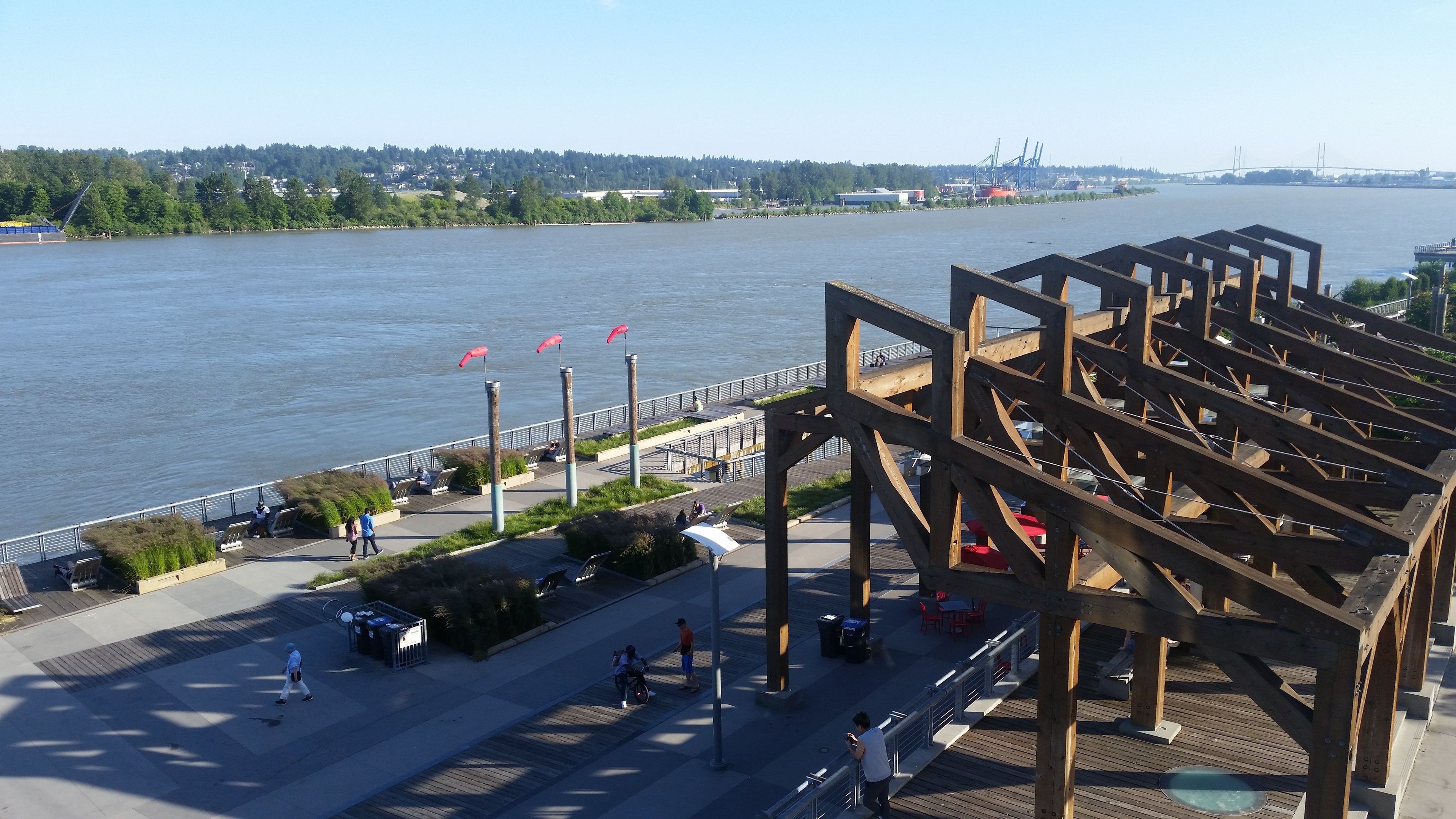 A riverside walkway with wooden structures, seating areas, and a view of the river and distant bridge.