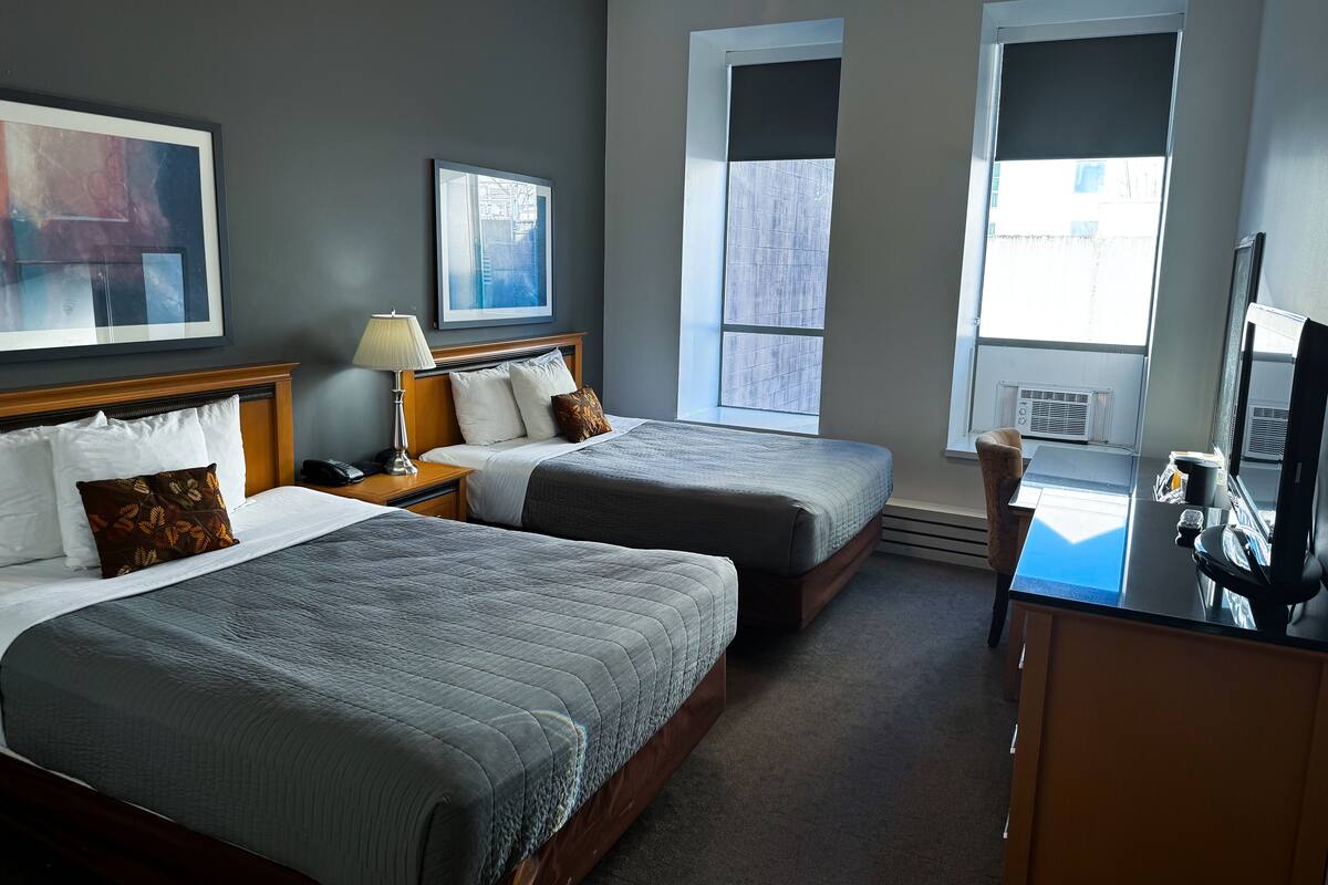 A hotel room with two beds, a desk, a chair, a ceiling fan, and large windows.