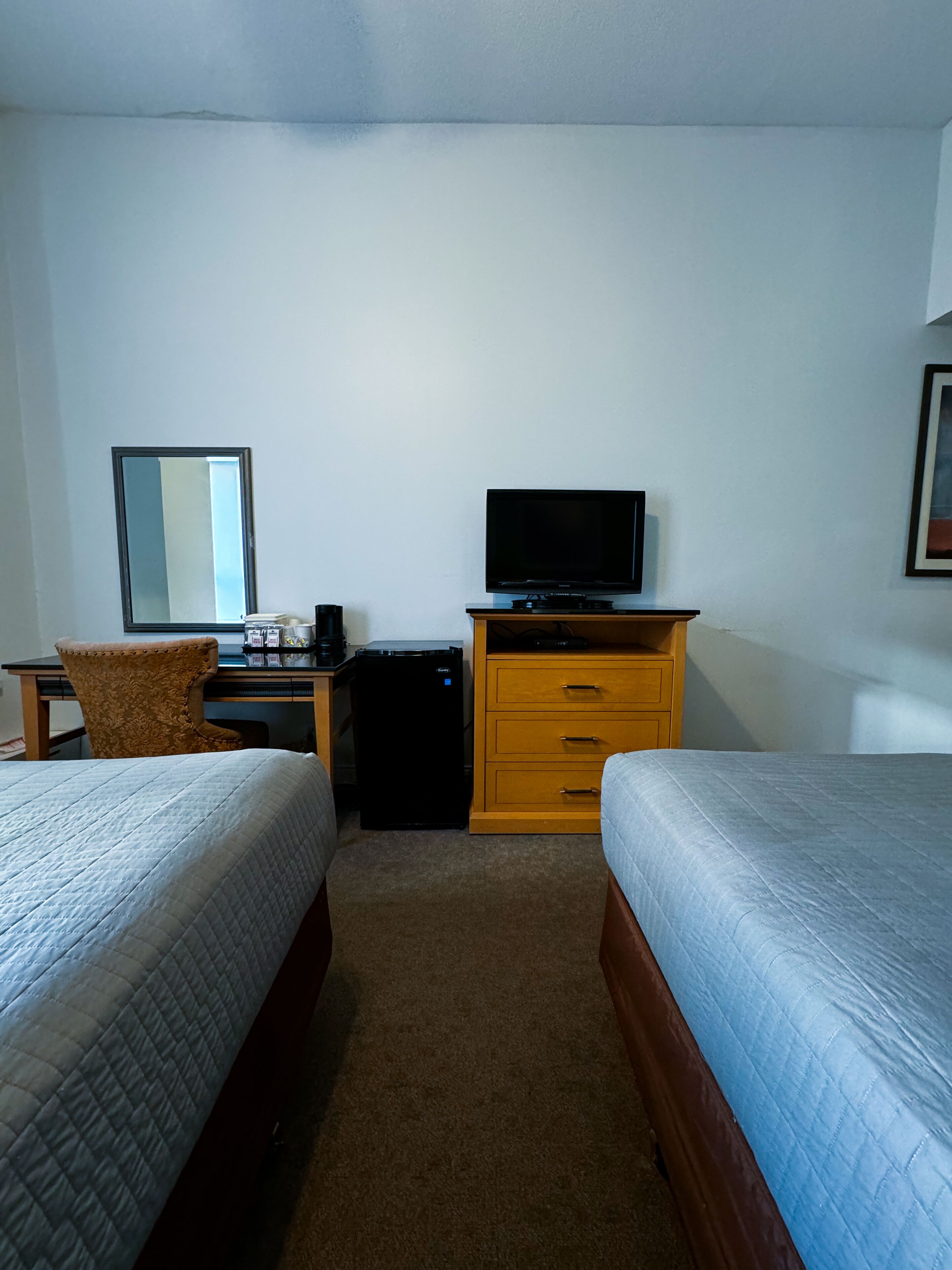 Una habitación de hotel con dos camas, un escritorio con computadora, televisor y un refrigerador pequeño.