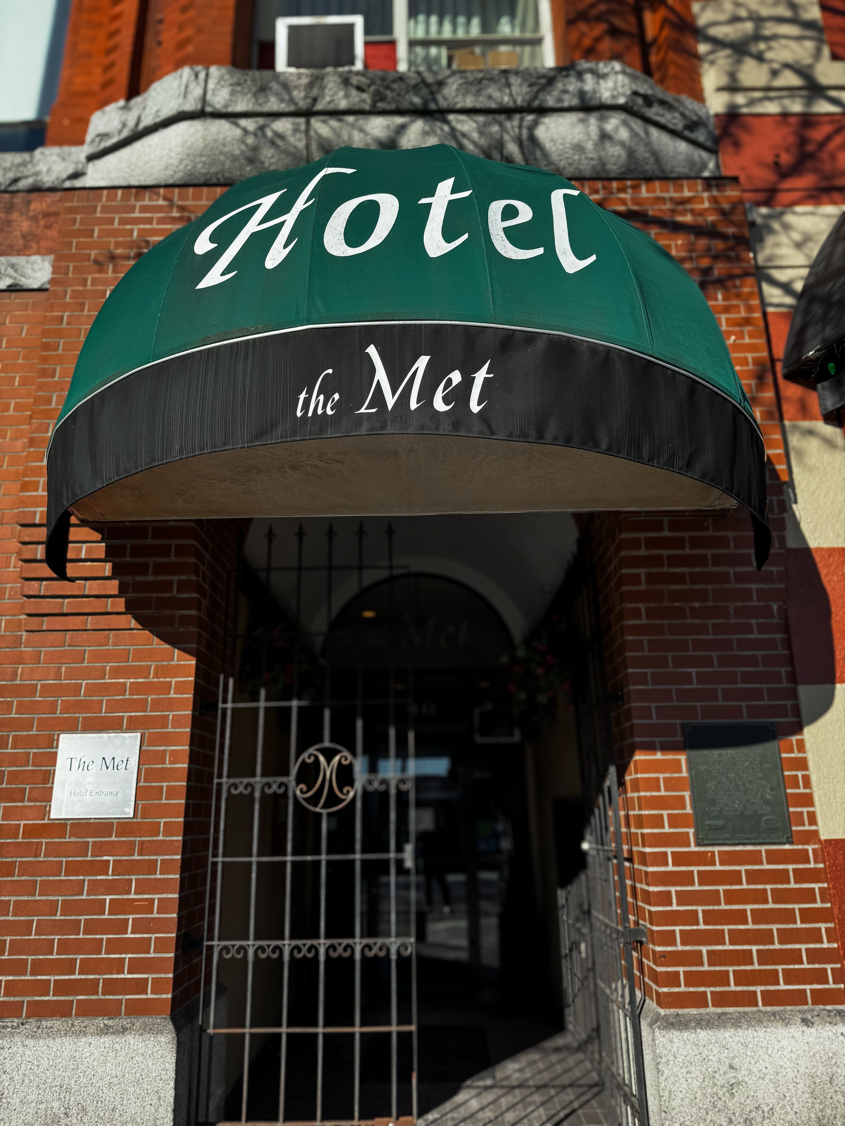Un toldo verde y negro con la inscripción "Hotel the Met", sobre una entrada de ladrillo con una reja metálica y un cartel que dice "Entrada al Hotel the Met".