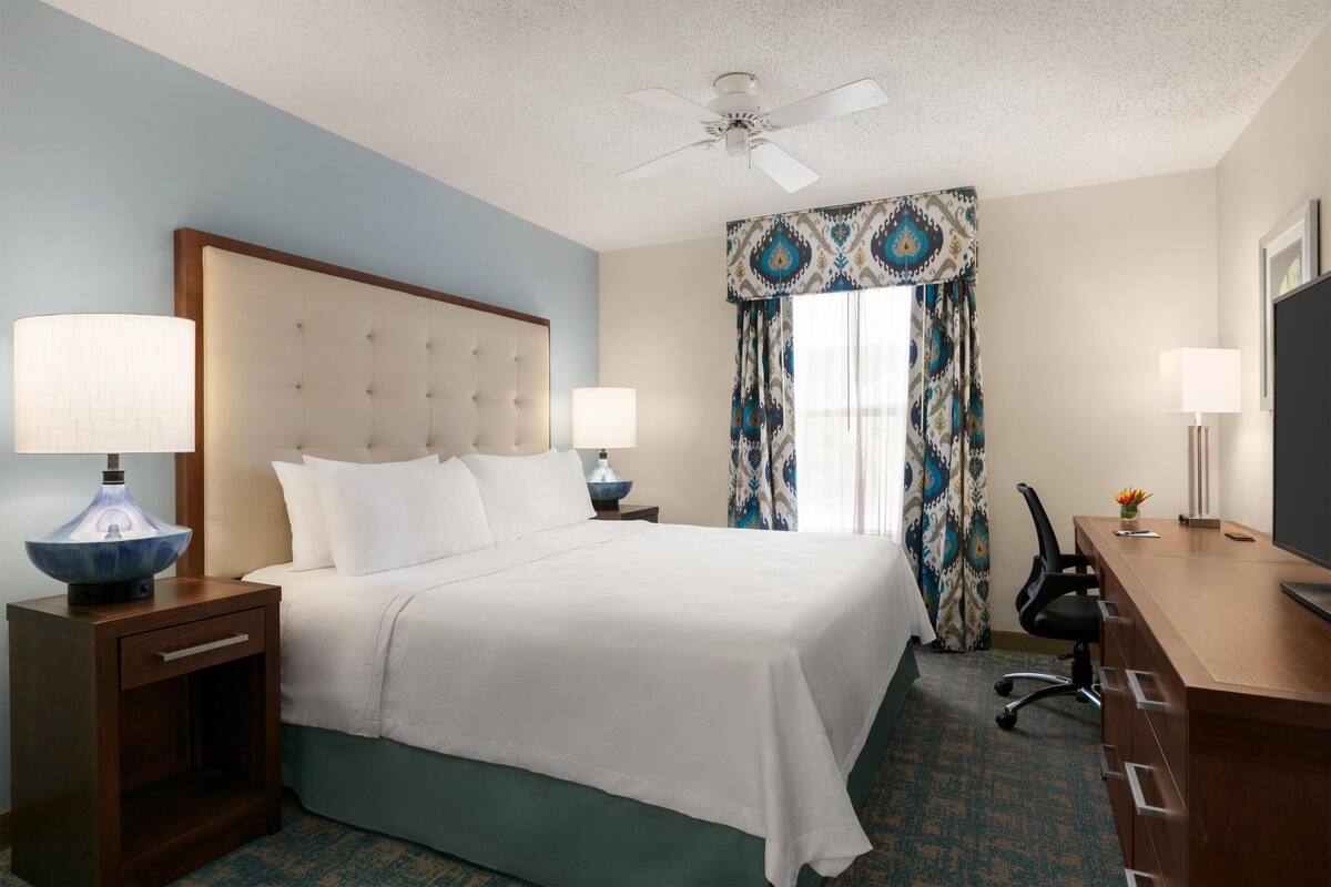 A hotel room with a bed, two bedside lamps, a desk with a chair, a television, and a ceiling fan.
