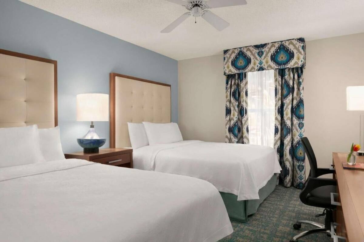A hotel room with two beds, a ceiling fan, a desk with a chair, and a window with patterned curtains.