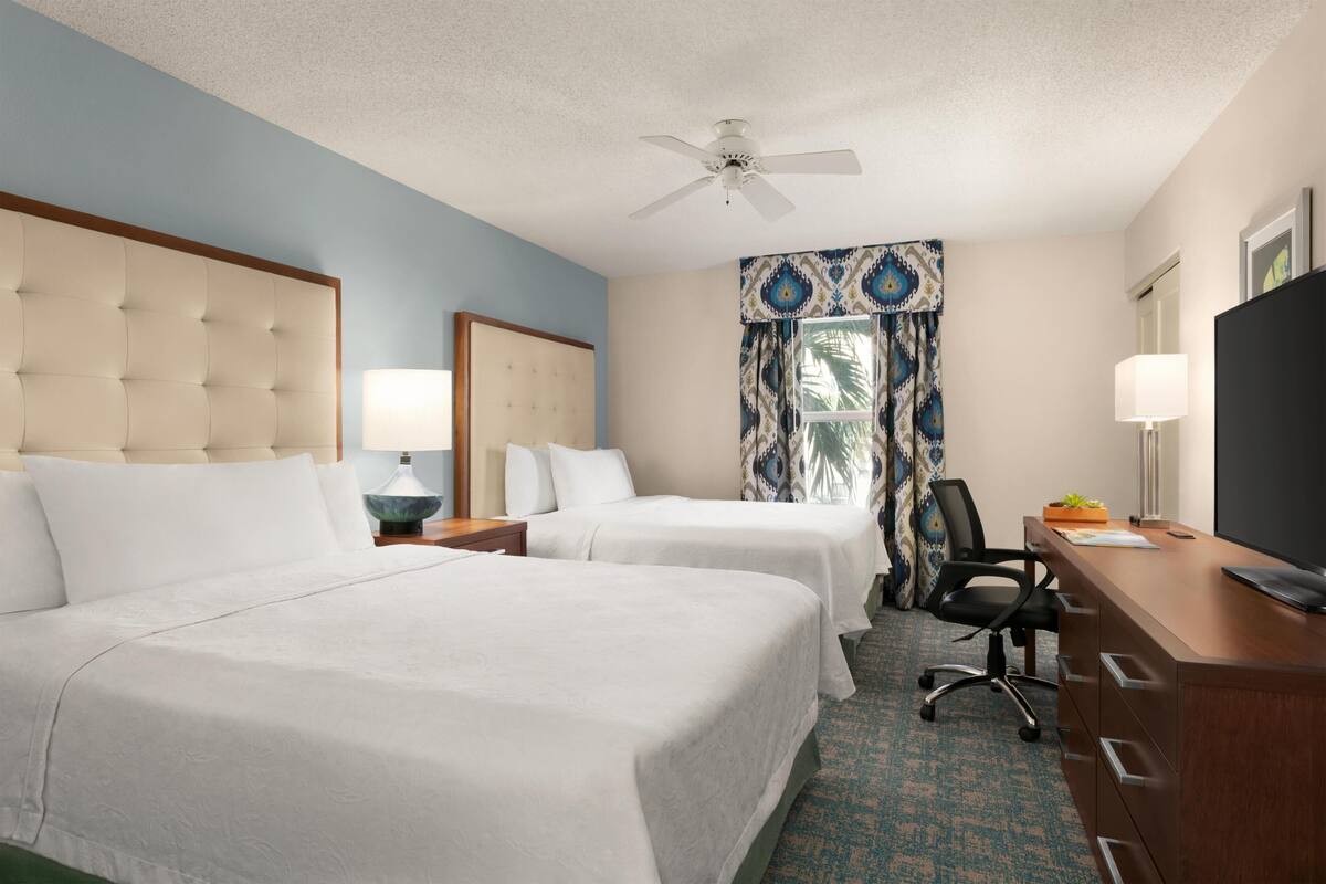 A hotel room with two beds, a desk, a chair, a television, a ceiling fan, and a window with patterned curtains.