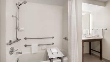 A bathroom with a bathtub, shower, handrails, a small table with a towel, and a sink with a mirror.