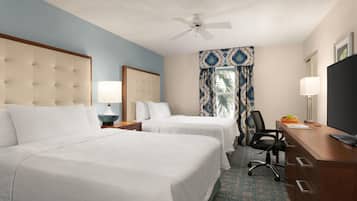 A hotel room with two beds, a desk, a chair, a television, a ceiling fan, and a window with patterned curtains.