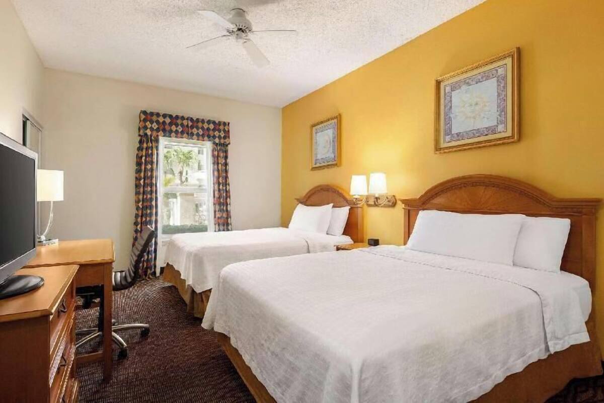 A hotel room with two beds, a desk with a TV, a ceiling fan, and two wall-mounted lamps.