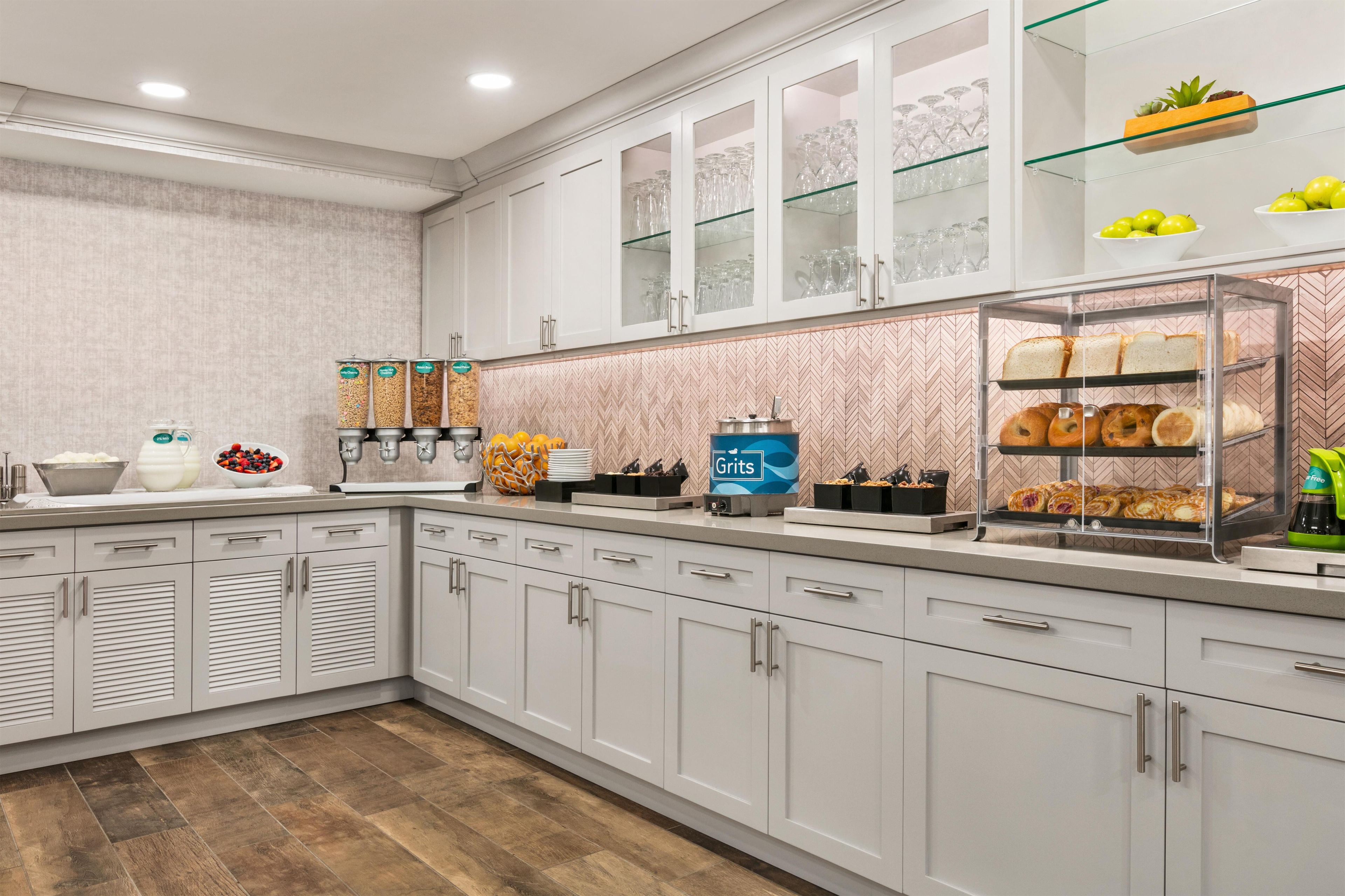 Uma cozinha moderna com armários brancos, painel de backsplash em padrão herringbone e uma vitrine de vidro com pão e doces.