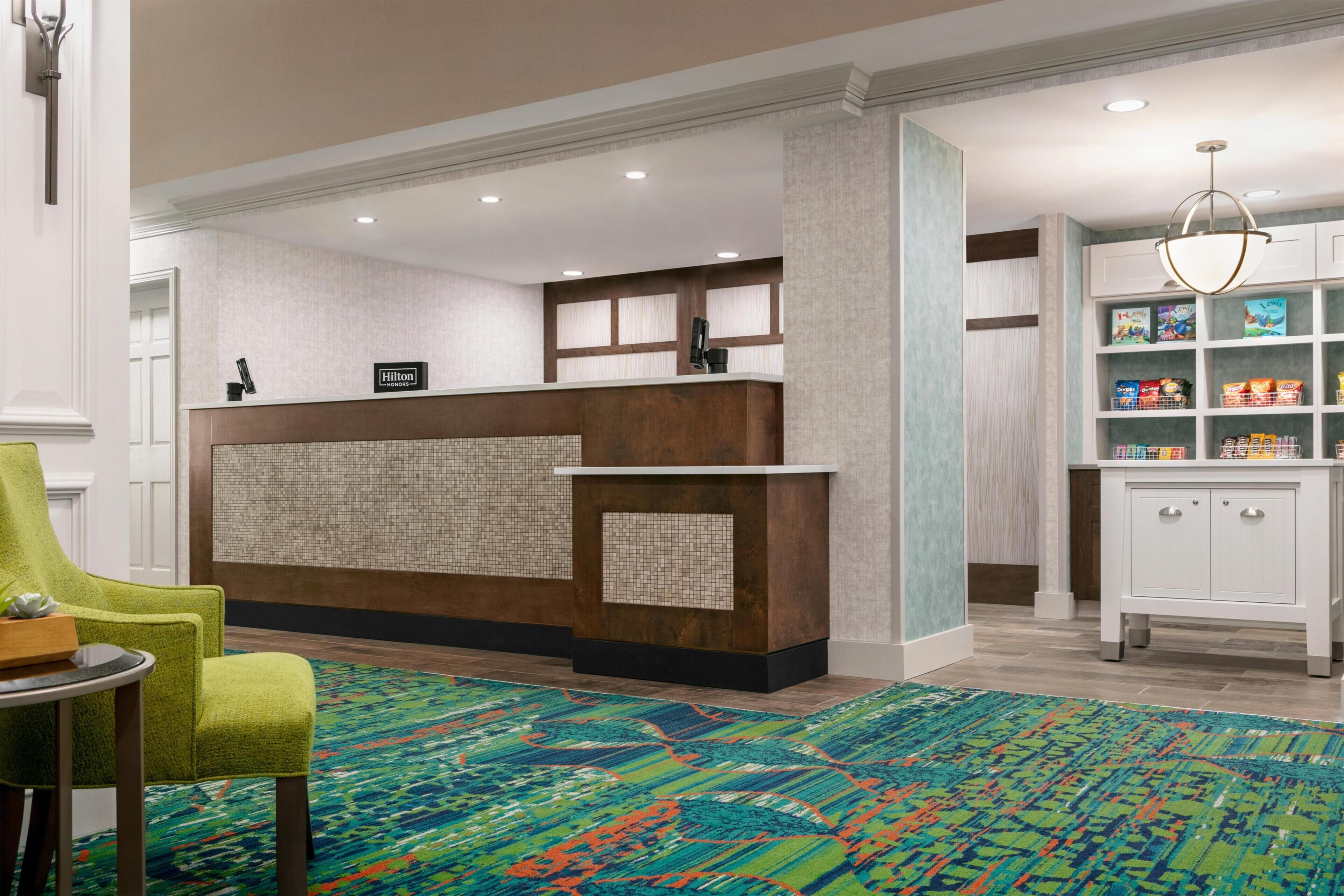 A hotel reception area with a wooden reception desk, a green armchair, a patterned rug, and a shelf with snacks.